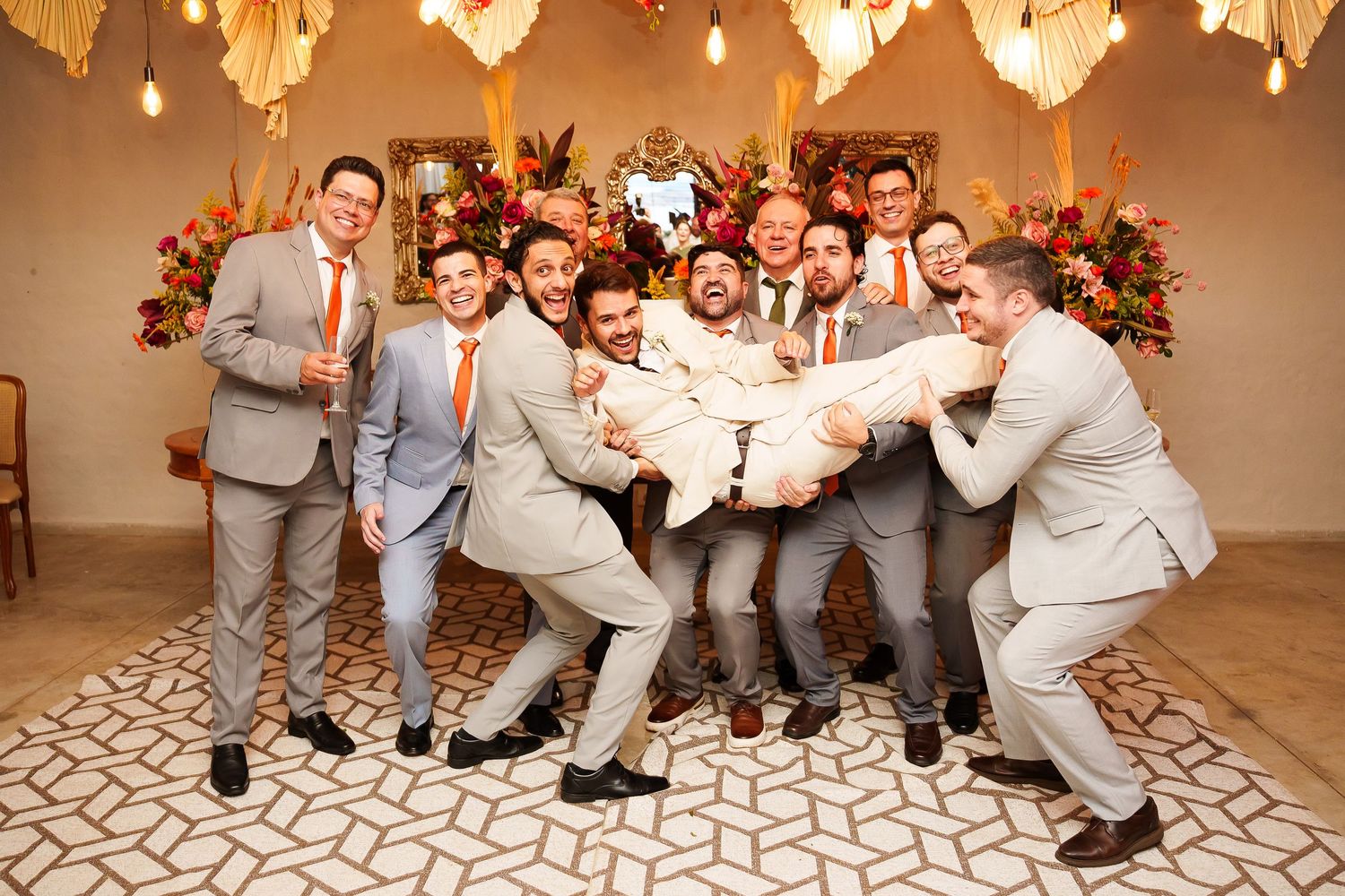 Groomsmen in gray suits with orange ties lift the groom during a fun wedding party photo indoors.