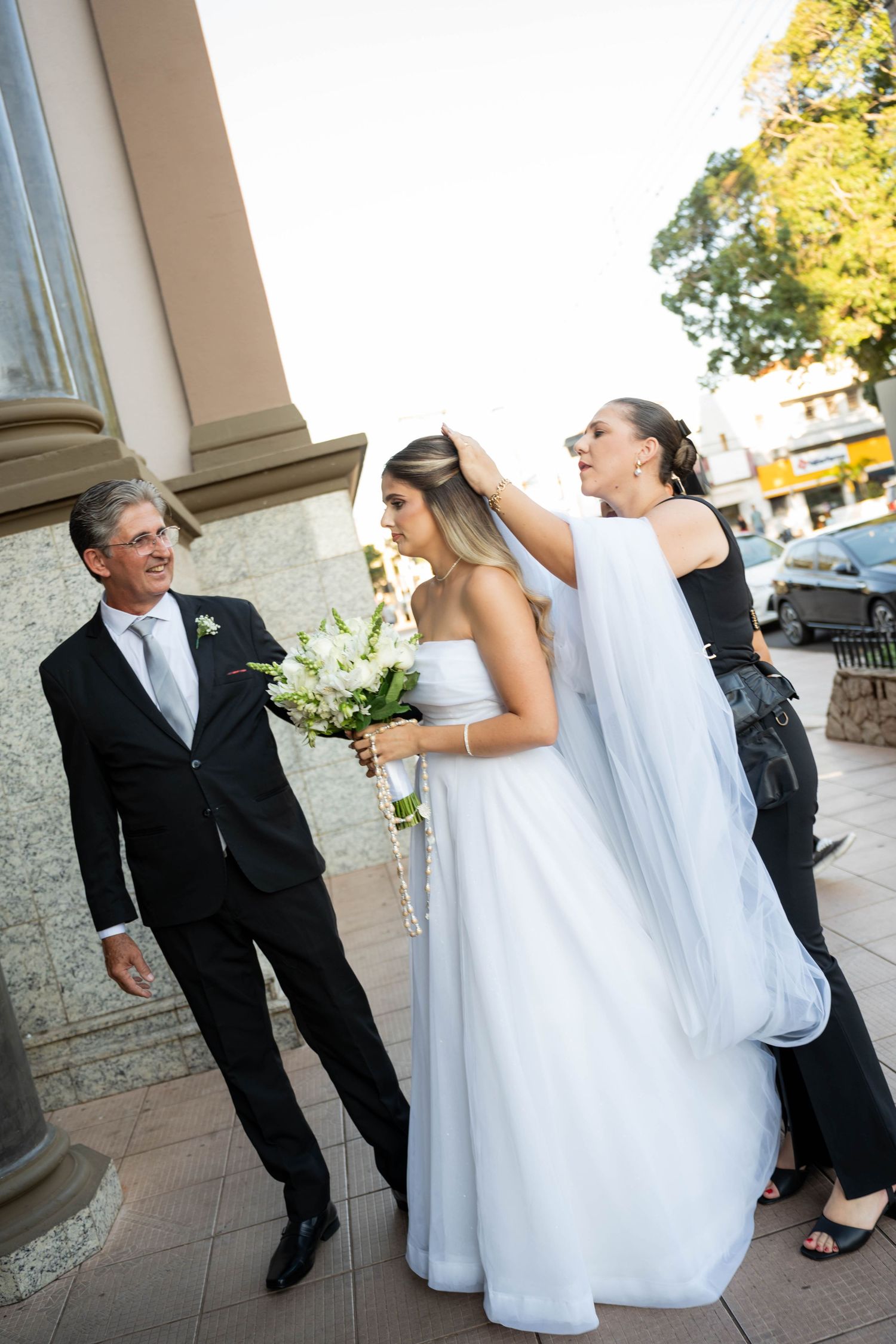 Bride in white gown holds bouquet while woman adjusts veil, man in suit smiles nearby outside church.