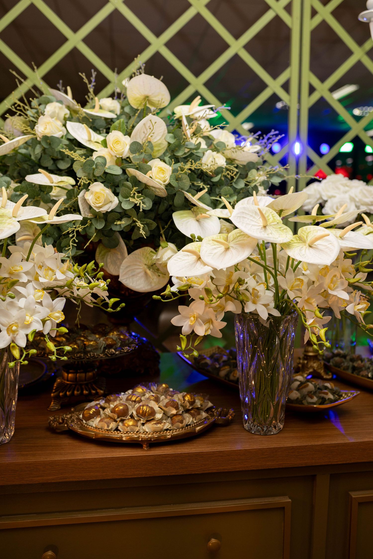 Elegant white anthurium and rose floral arrangements in crystal vases on a wooden table at a formal event.
