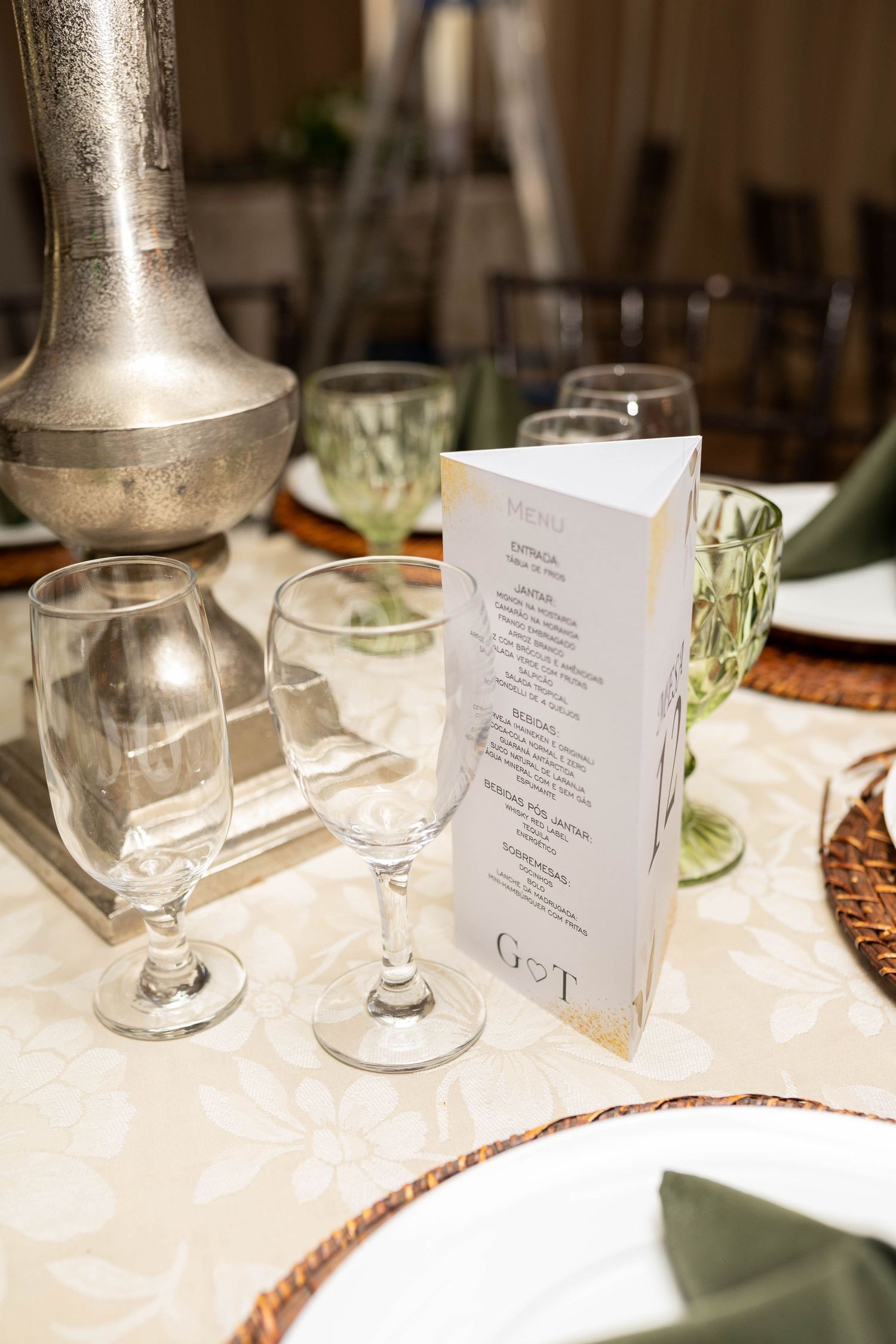 Elegant wedding table setting with menu card, crystal glasses, green goblets, and wicker charger plates on floral tablecloth.