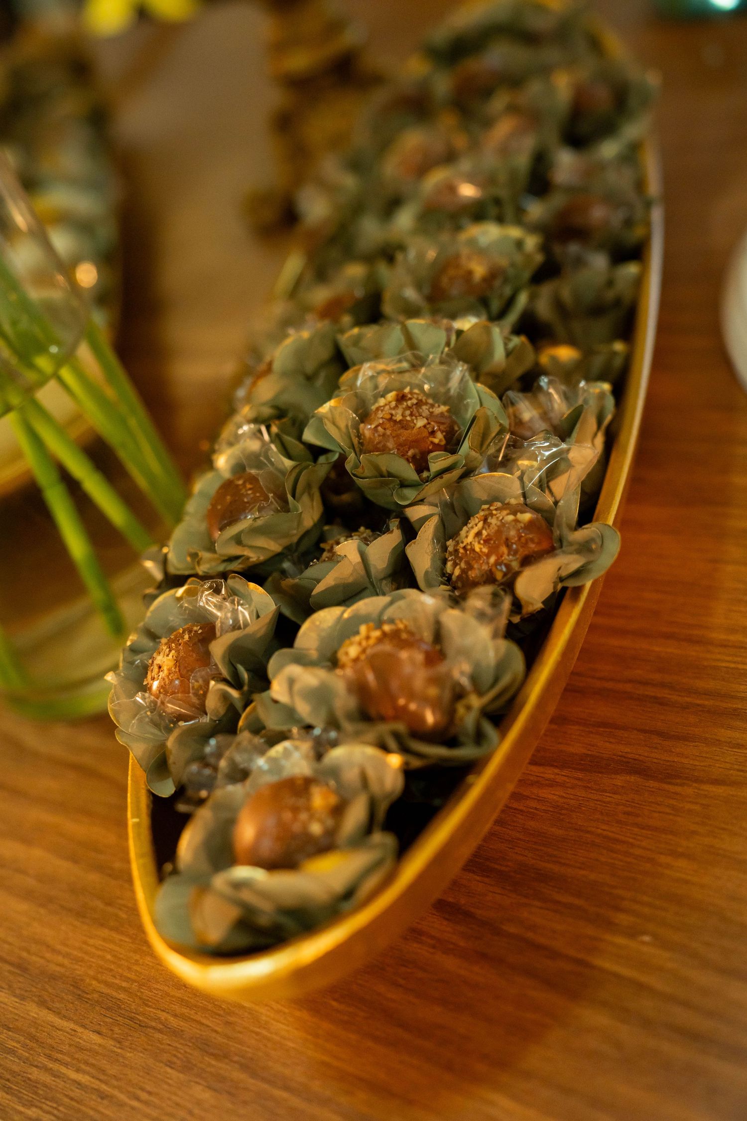 Elegant gold boat dish filled with Ferrero Rocher chocolates wrapped in green foil flowers on a wooden table.