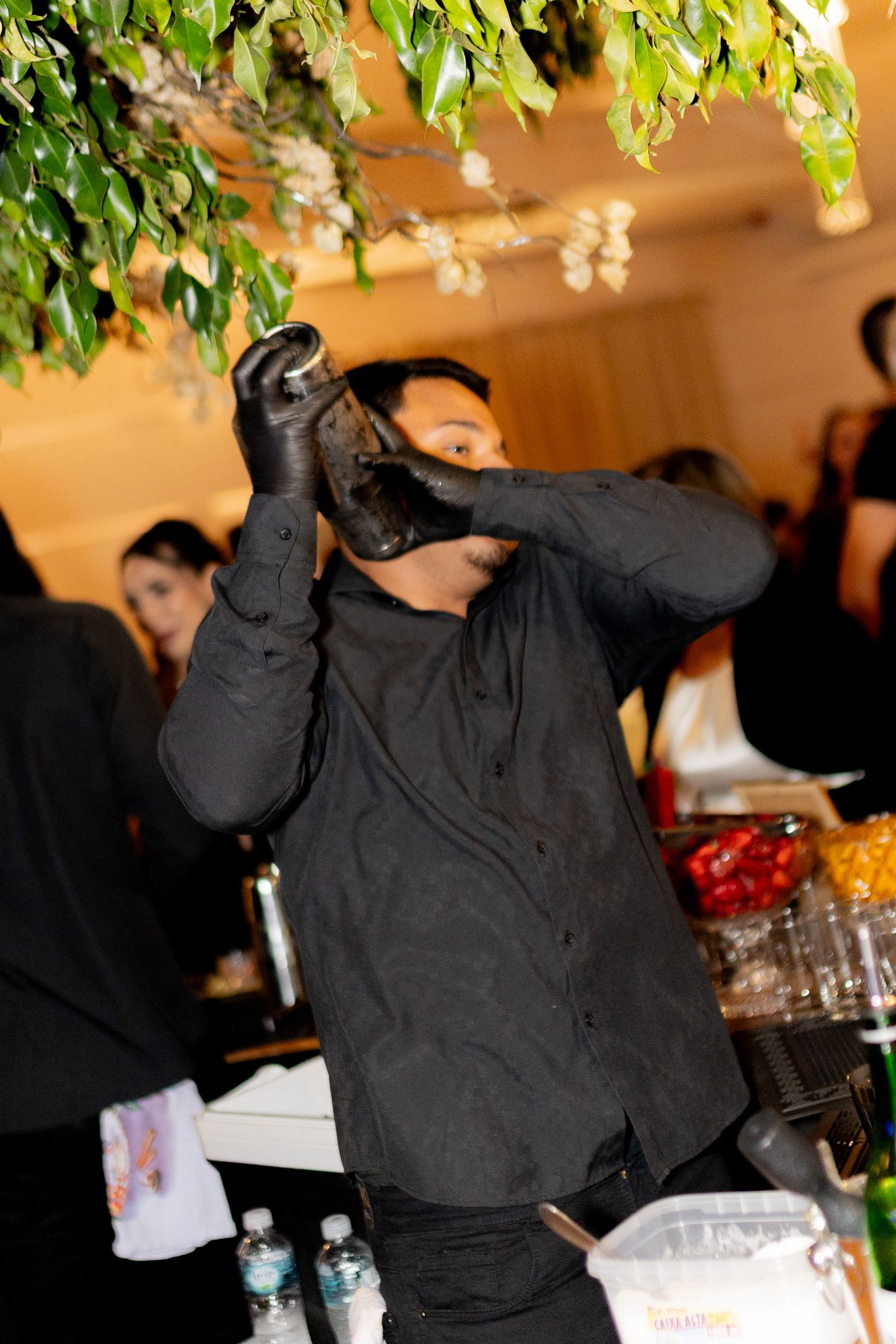 Bartender in black outfit and gloves mixing a cocktail at an indoor event bar.