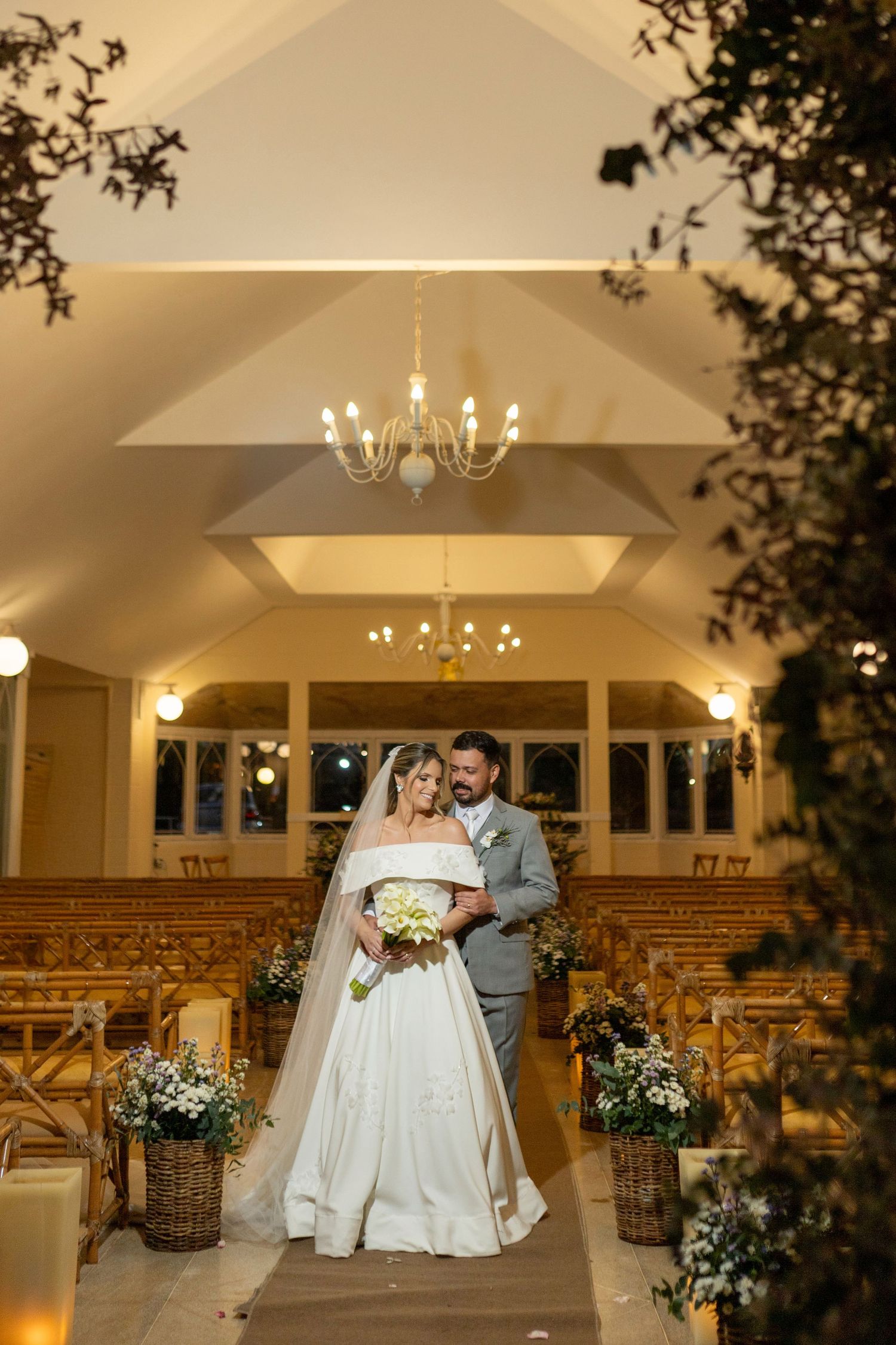 Newlyweds pose in chandelier-lit chapel aisle, bride holding bouquet in elegant ball gown with veil.