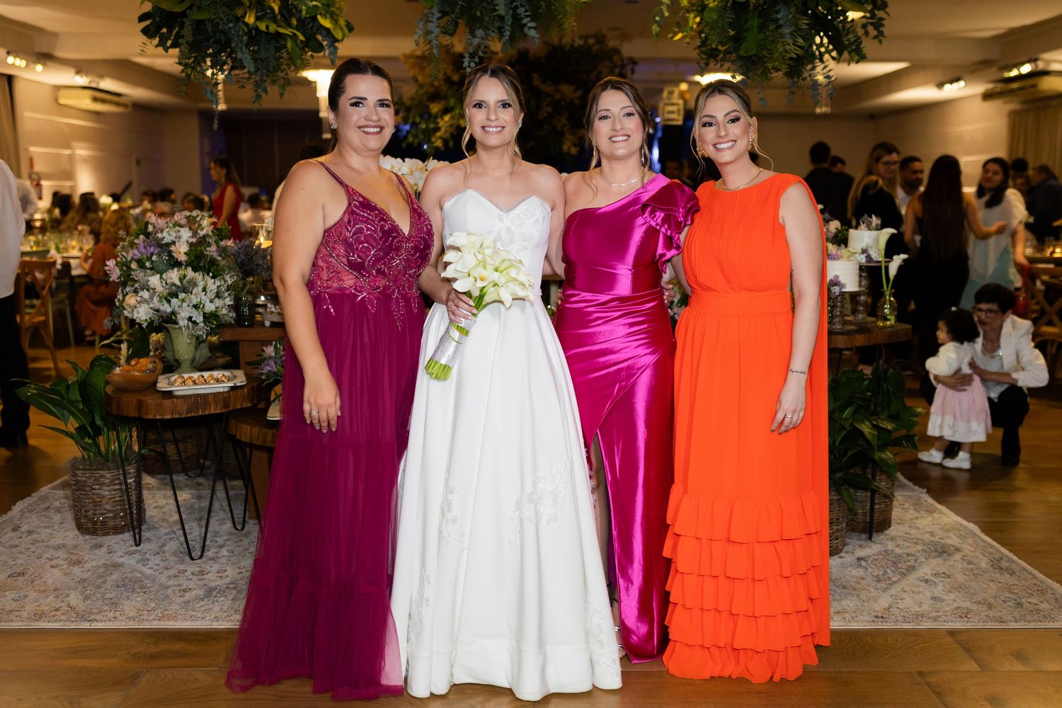 Bride in white gown poses with three women in colorful dresses at an elegant wedding reception venue.