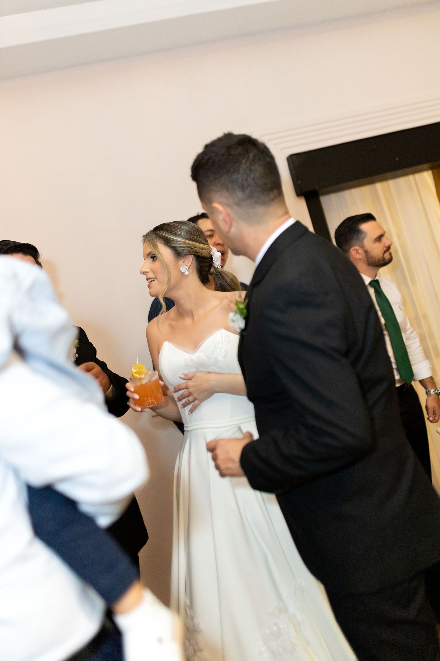 Smiling bride in white gown walks with groom in black suit at wedding reception surrounded by guests.