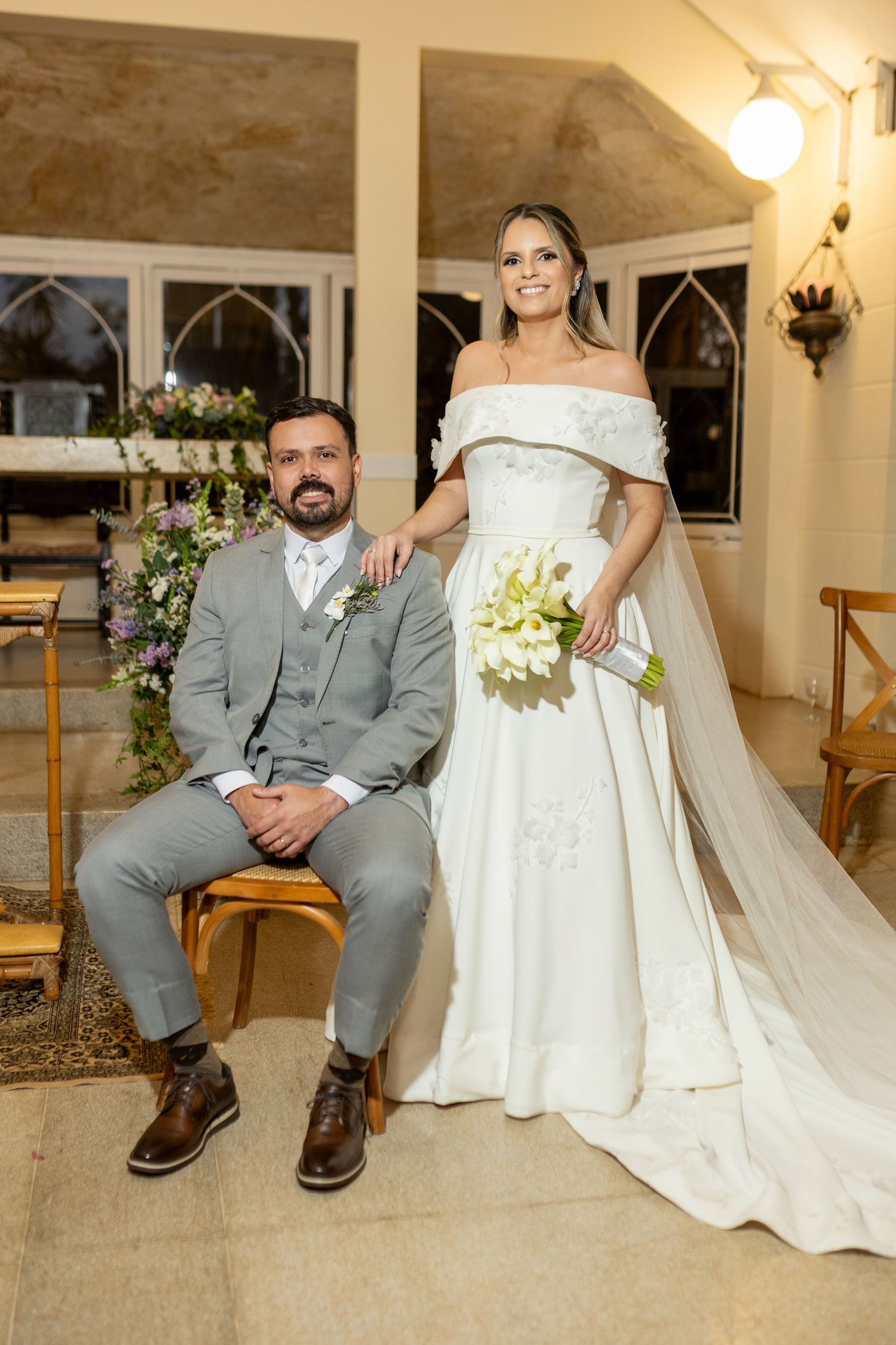Bride in off-shoulder white gown with bouquet stands beside groom in gray suit seated in elegant church setting.