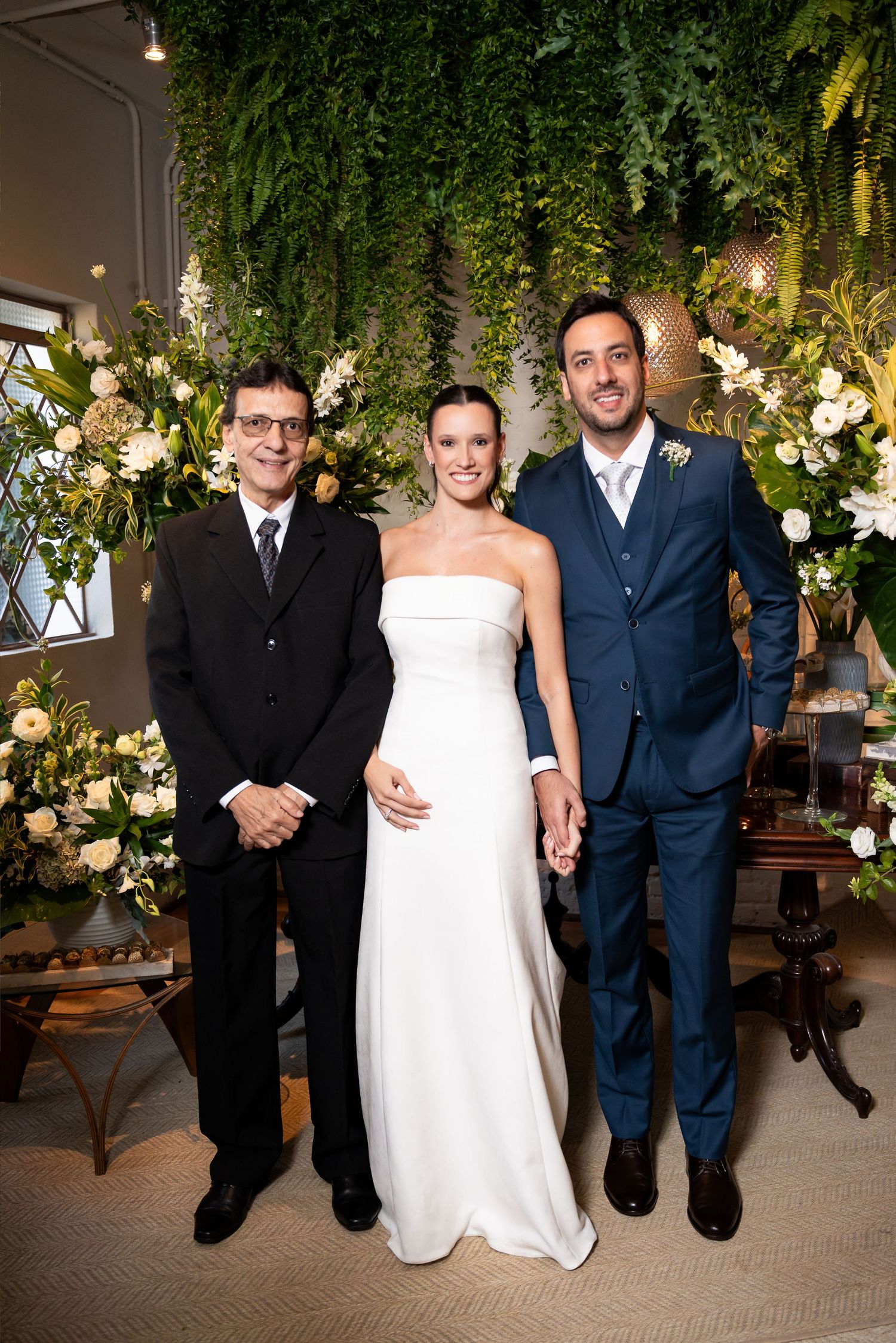 Bride in white strapless gown stands with groom in navy suit and officiant in black suit at floral wedding ceremony.