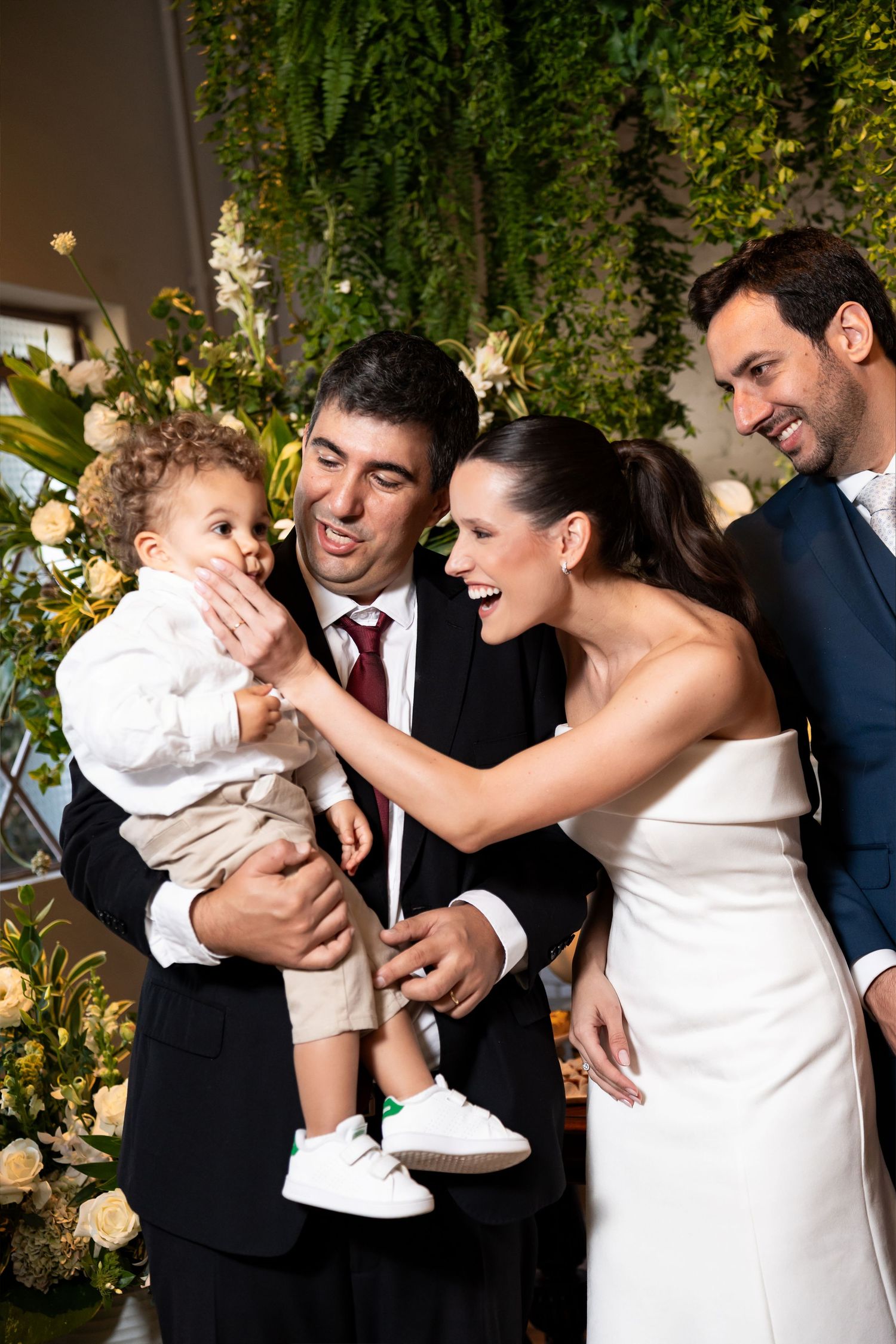 Bride and groom in wedding attire joyfully interact with a baby boy amid floral decorations.