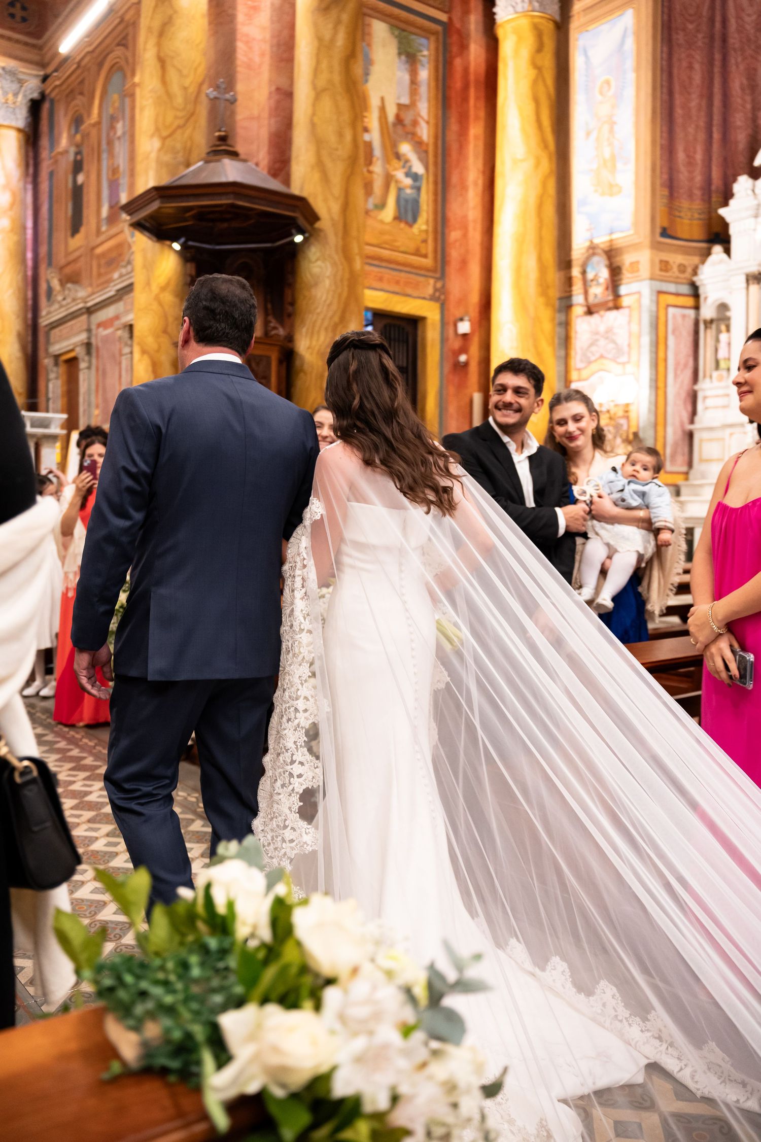 Bride and groom walking down the aisle in an ornate church, with guests smiling and a flowing white veil trailing behind.