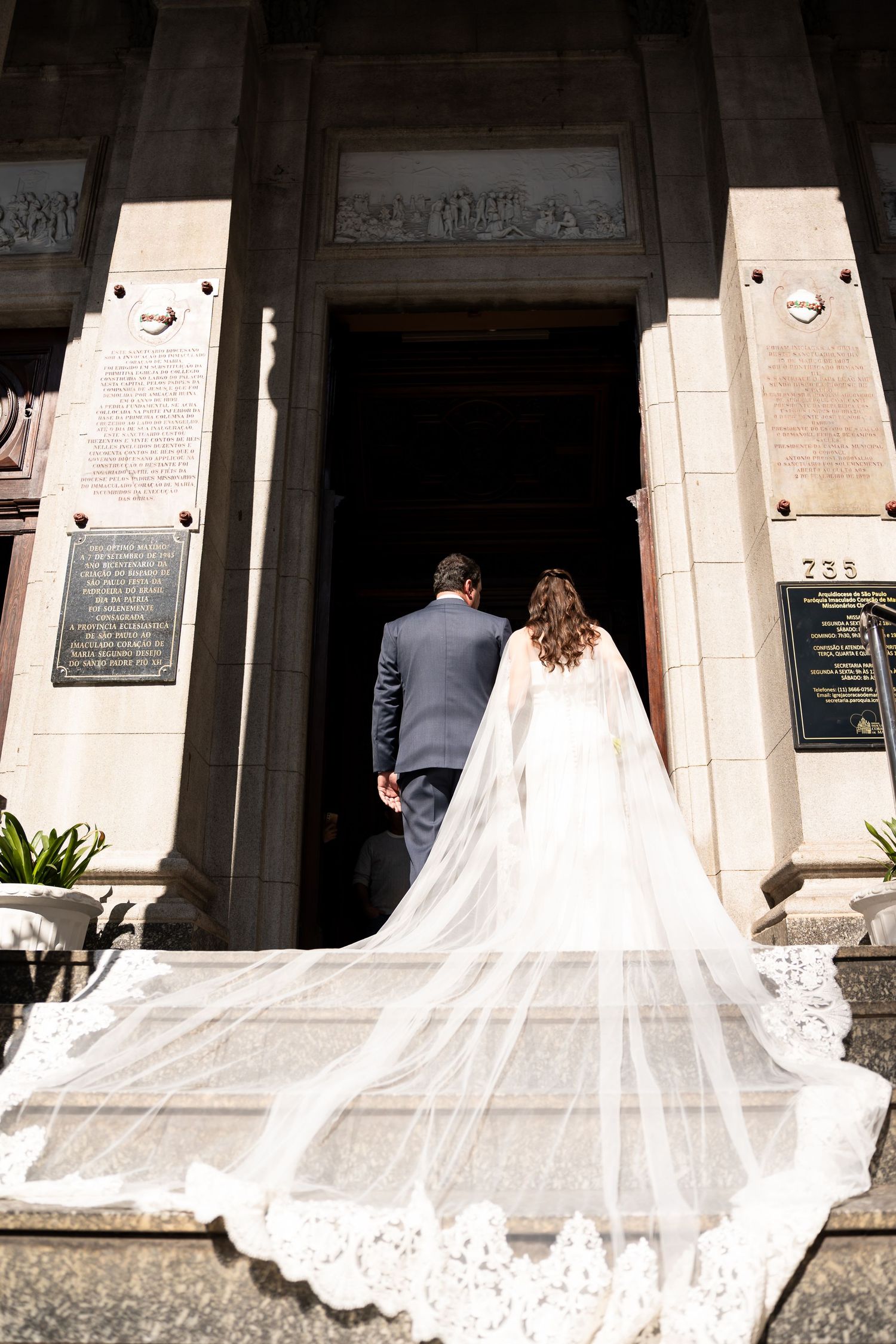 Bride and groom walking into a church, with a long lace cathedral veil trailing behind on the stone steps.