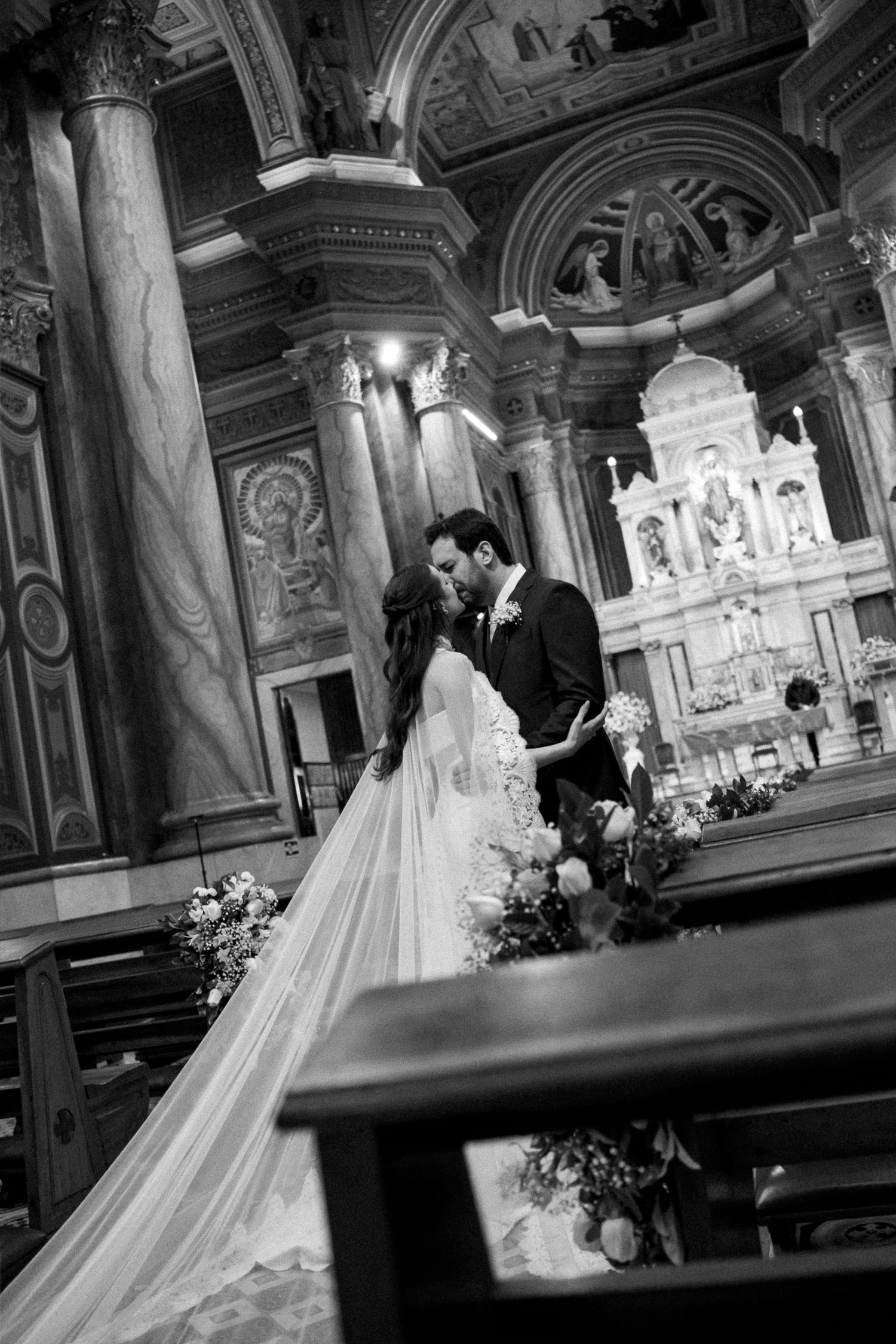 Black and white photo of bride and groom sharing a kiss at the altar of an ornate church during their wedding ceremony.
