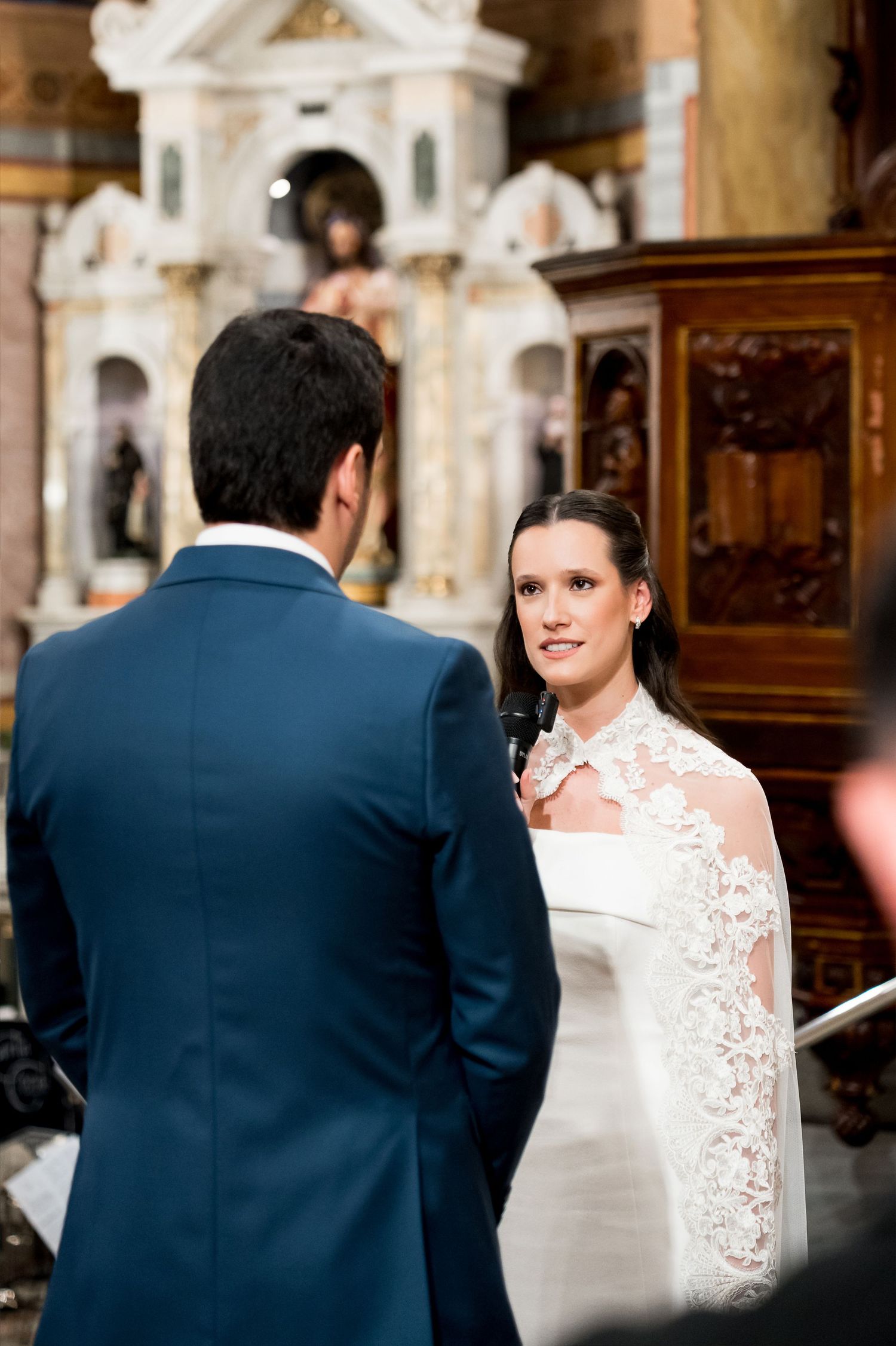 Bride in lace wedding dress exchanging vows with groom in blue suit during church ceremony.