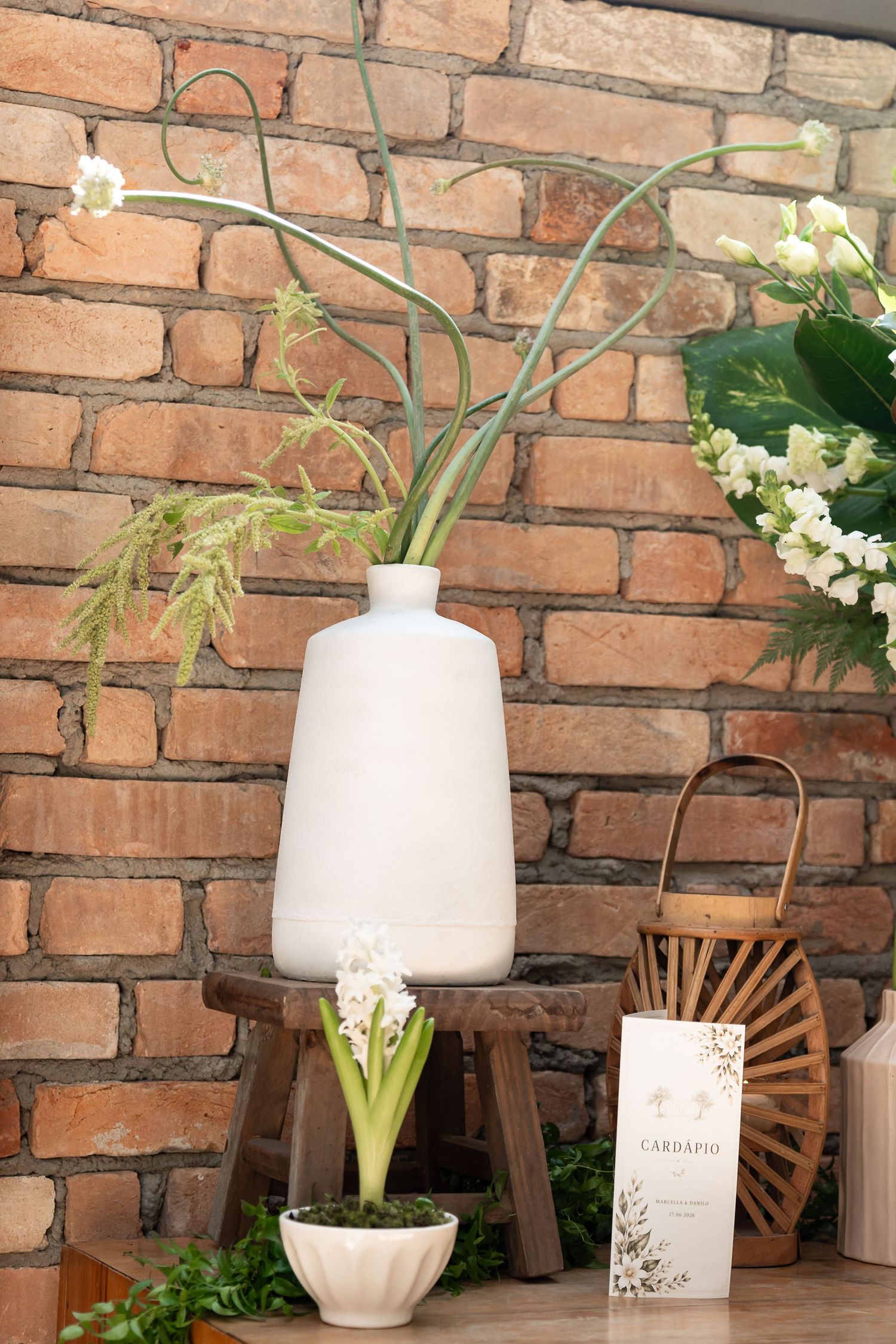 White ceramic vase with tall stems on wooden stool, hyacinth plant, and wicker basket against rustic brick wall.