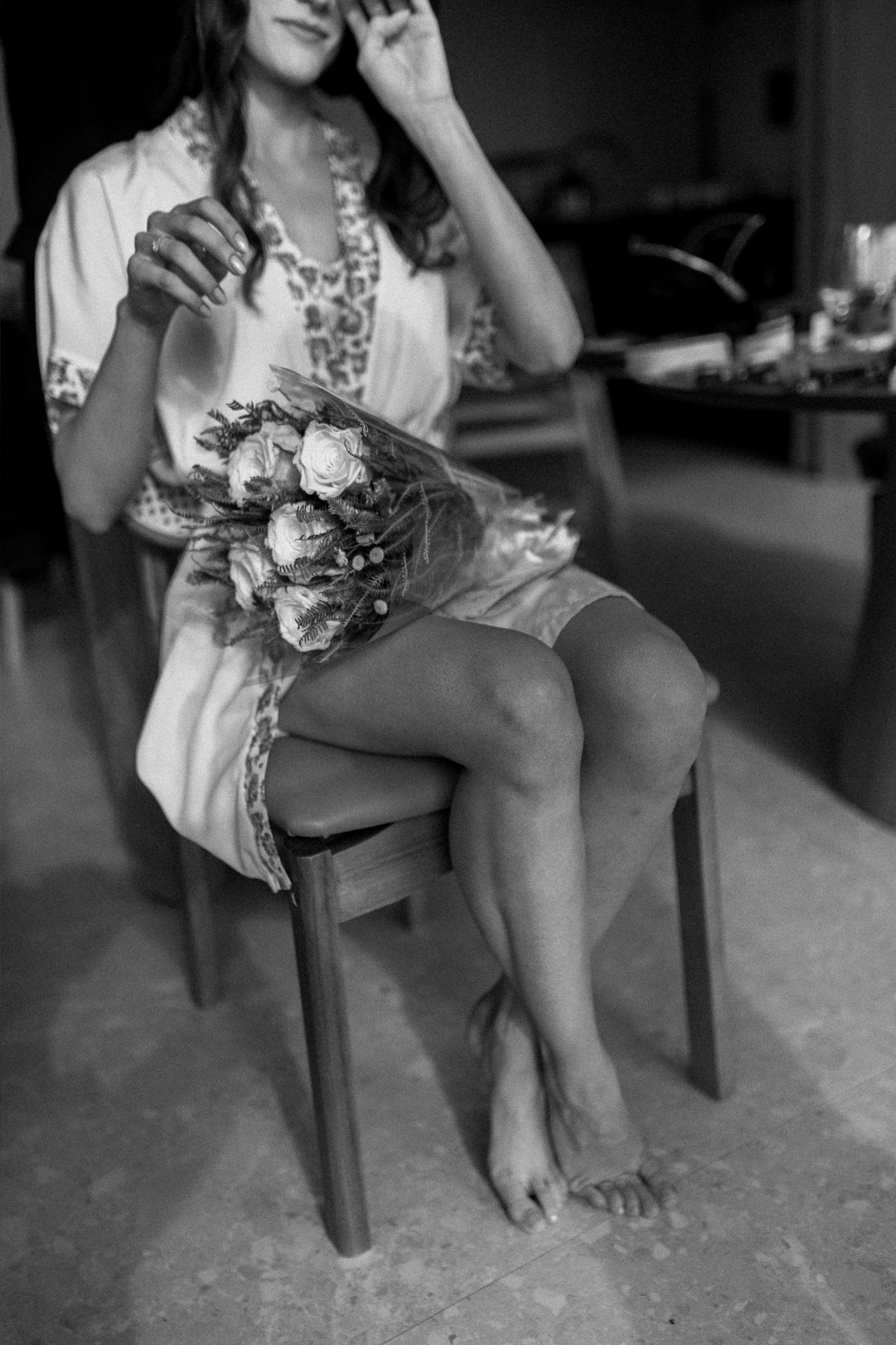Black and white photo of a bride in a robe holding a floral bouquet while sitting barefoot on a chair.