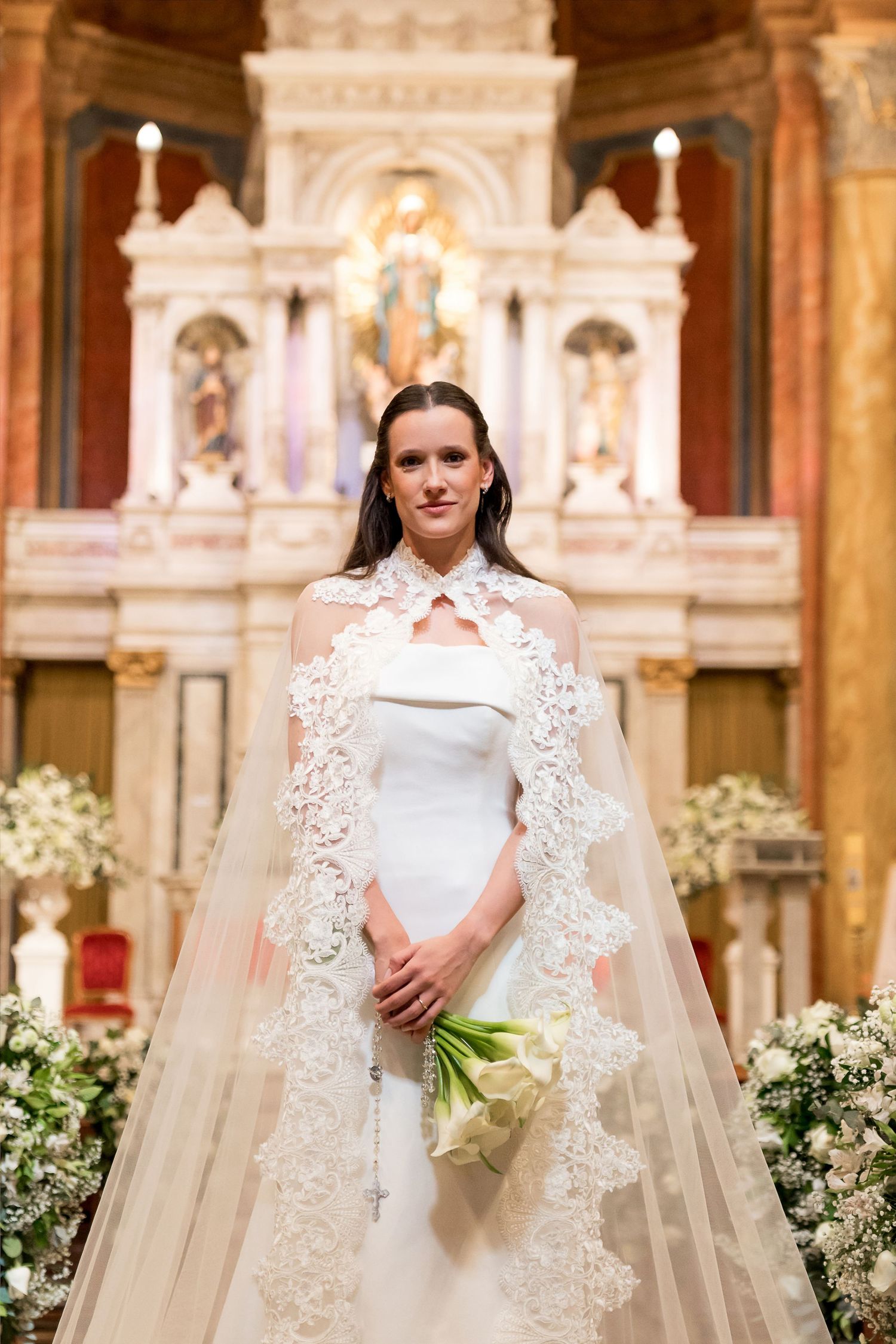 Bride in white gown with lace cape holds bouquet before ornate church altar adorned with floral arrangements.