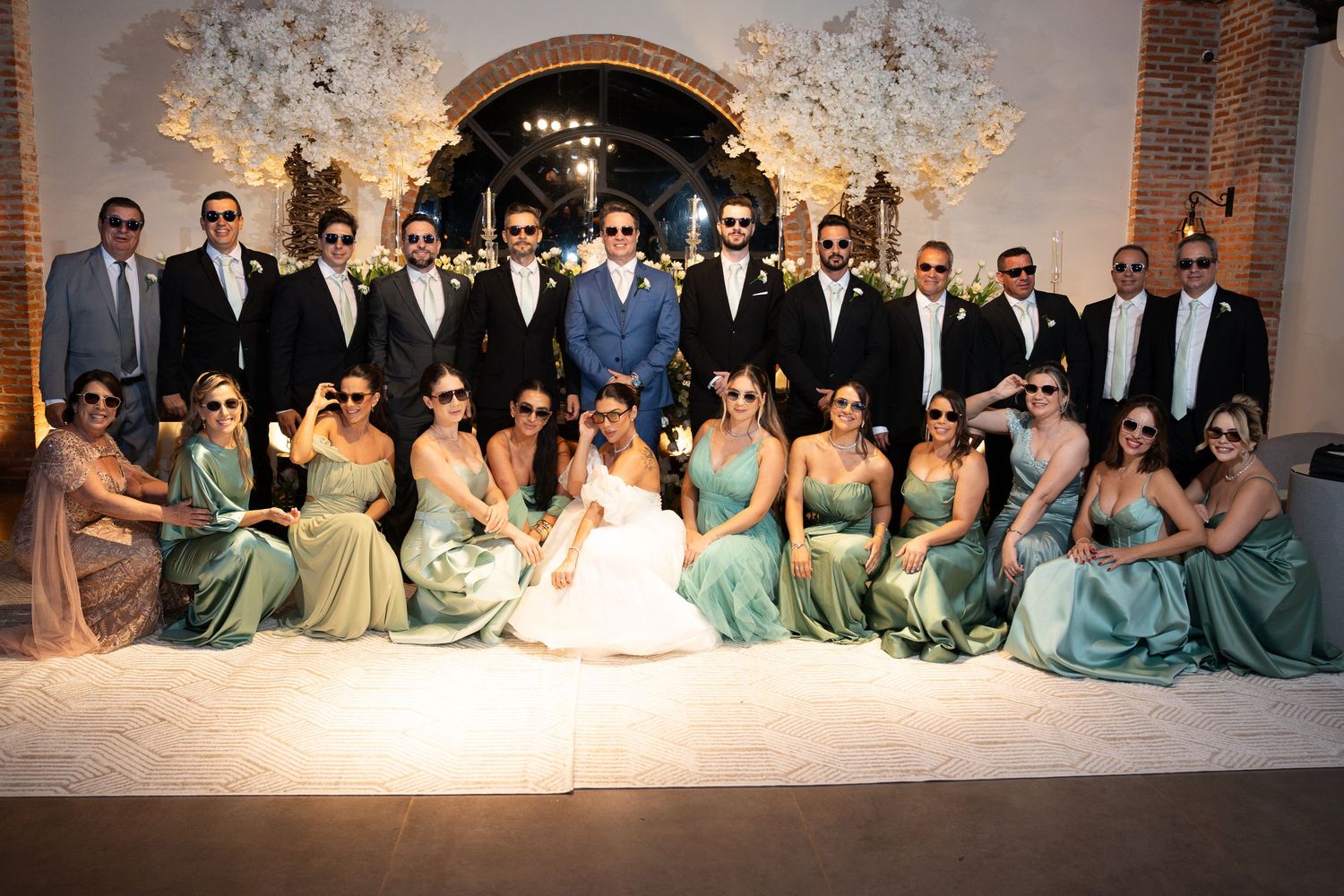 Wedding party poses together, bridesmaids in sage green gowns and groomsmen in black suits with sunglasses at elegant venue.