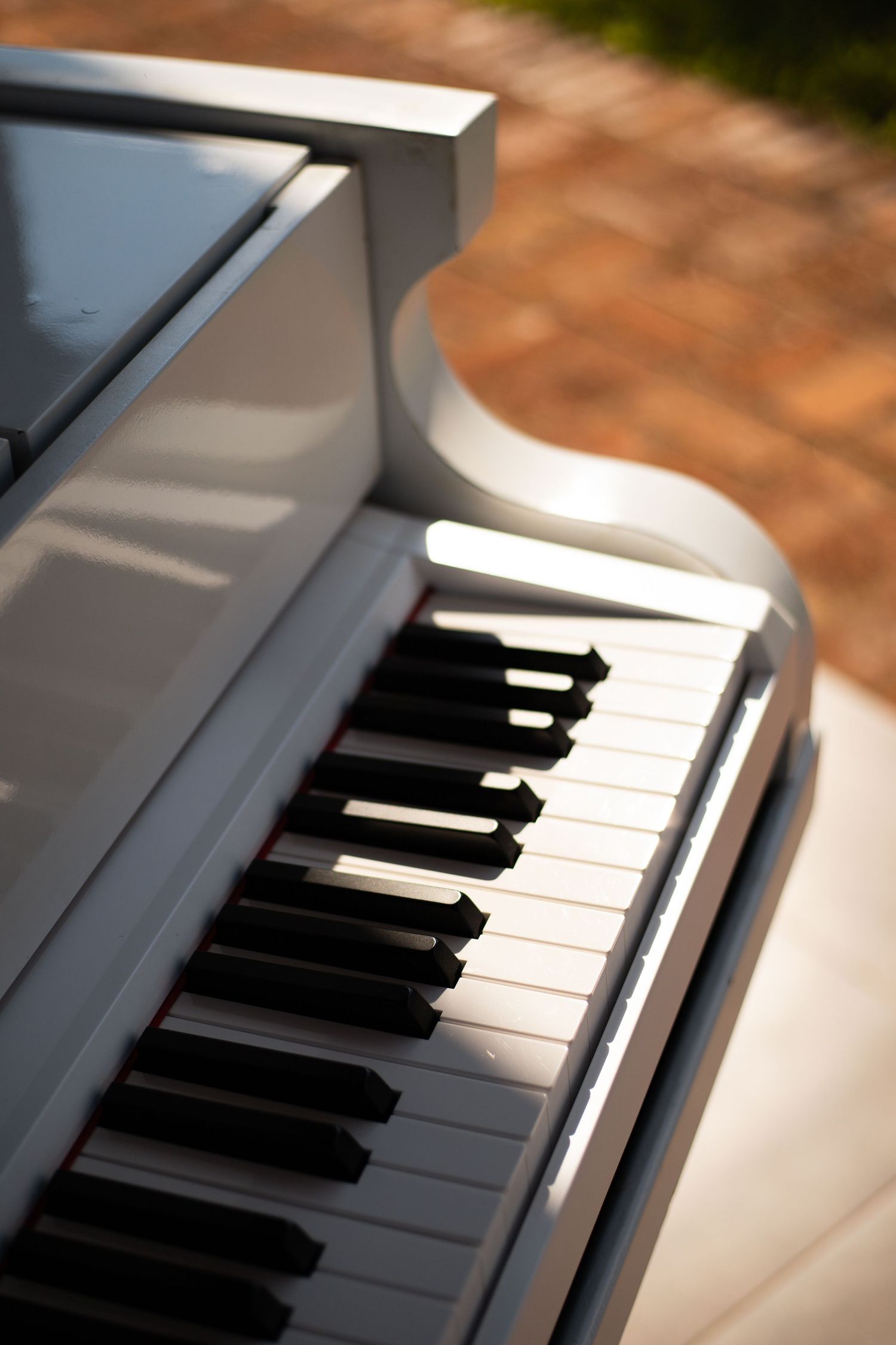White grand piano with black and white keys photographed outdoors in warm sunlight on a brick patio.