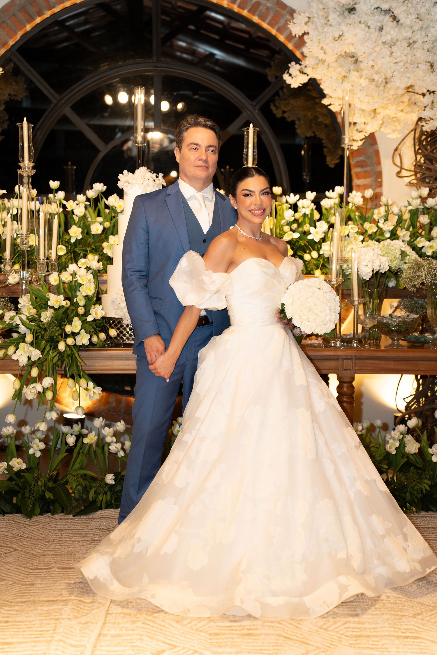 Bride in a white ballgown and groom in a blue suit pose together surrounded by white floral arrangements at their wedding.