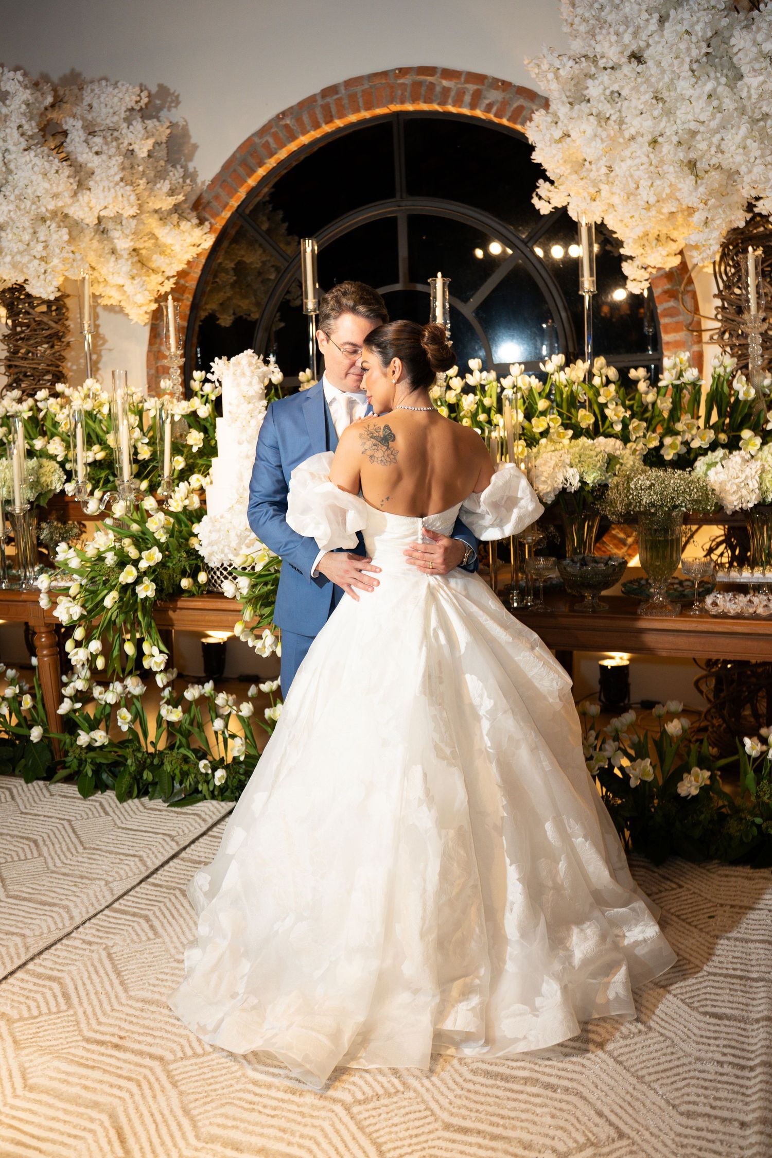 Bride in off-shoulder ball gown embraces groom in blue suit surrounded by white floral arrangements at elegant wedding reception.