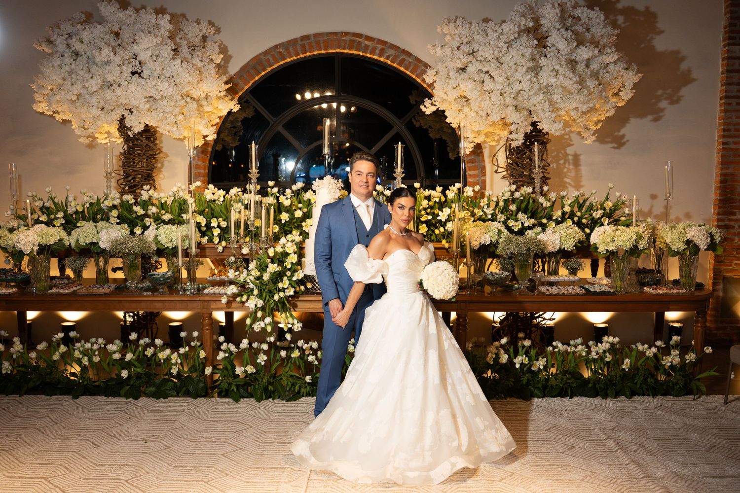 Elegant wedding couple posing before floral arch with white blooms and candlelit reception table in ornate venue.