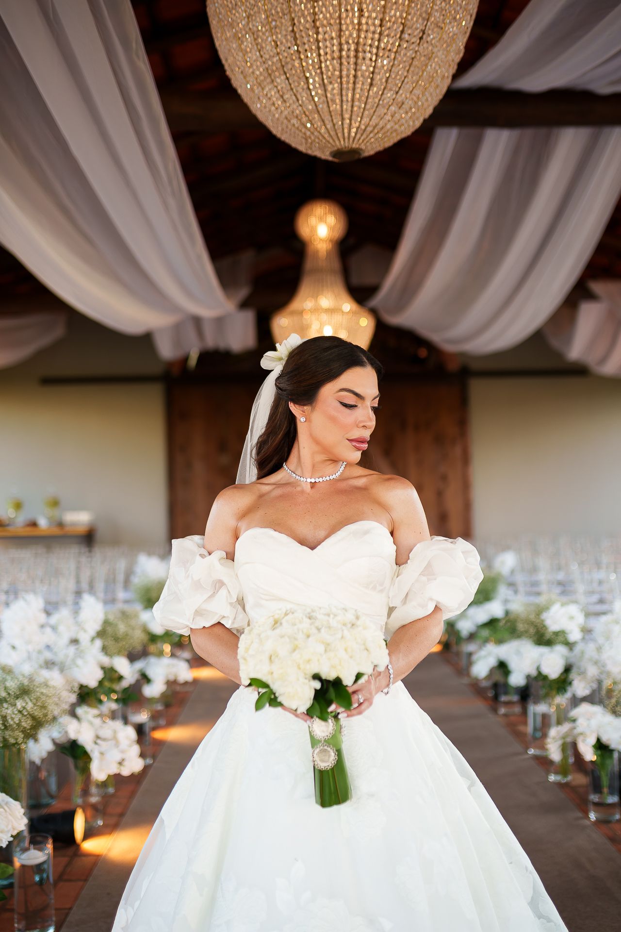 Elegant bride in off-shoulder white ballgown holding white bouquet stands in decorated wedding venue with chandeliers.