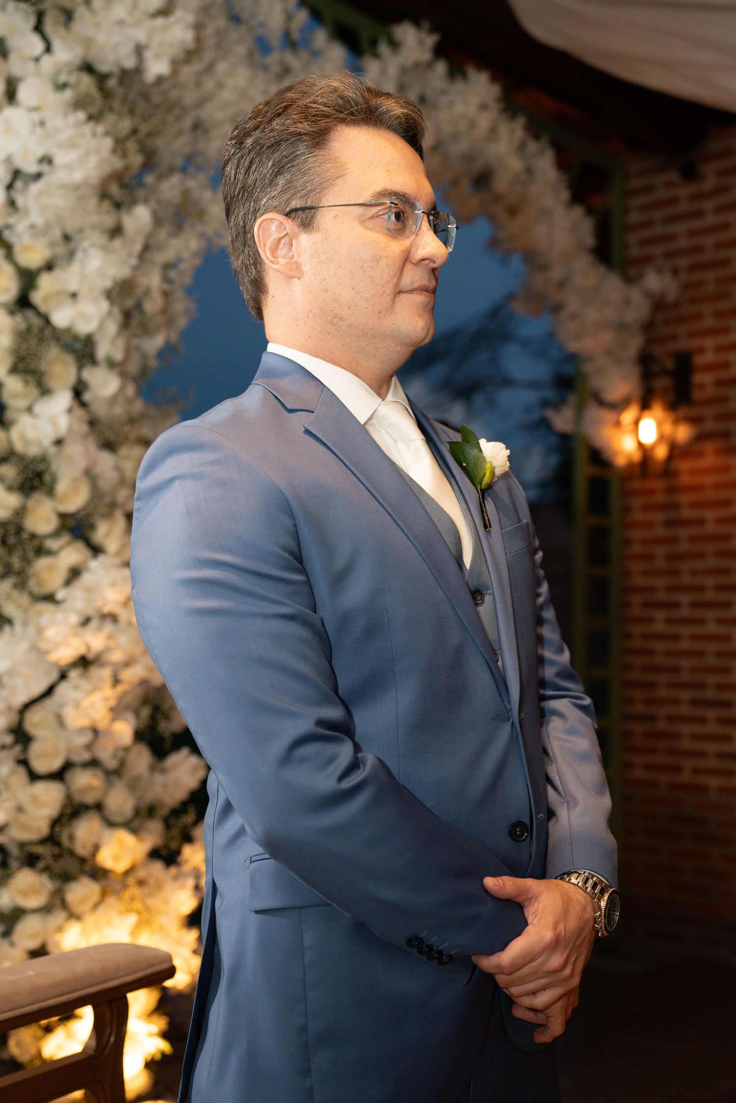 Groom in blue suit with boutonniere stands at wedding altar surrounded by white floral arrangements.
