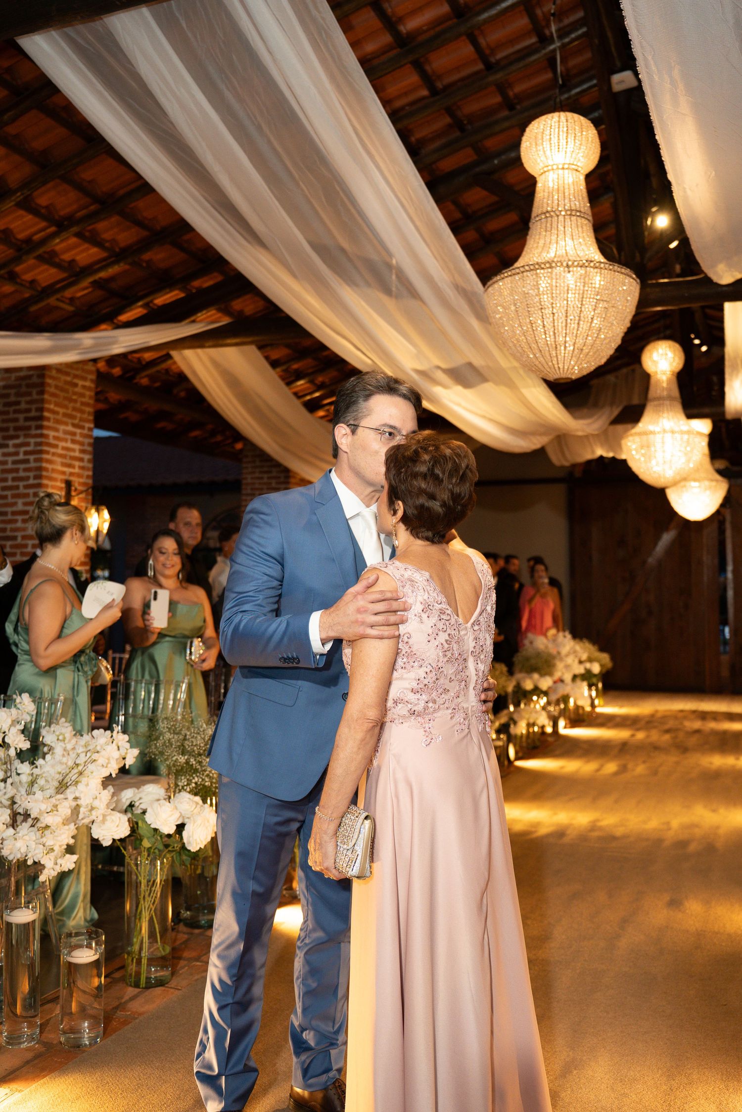 Groom in blue suit shares first dance with mother in pink gown at elegant wedding reception with chandeliers.