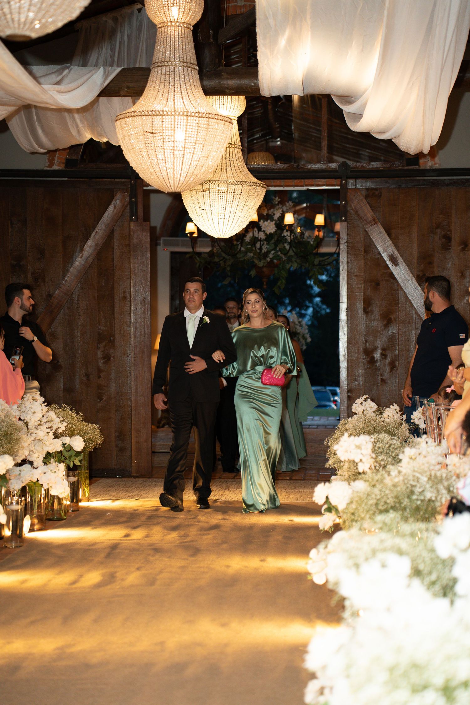 Couple walks down wedding aisle in elegant barn venue with chandeliers, woman in green gown, man in black tuxedo.