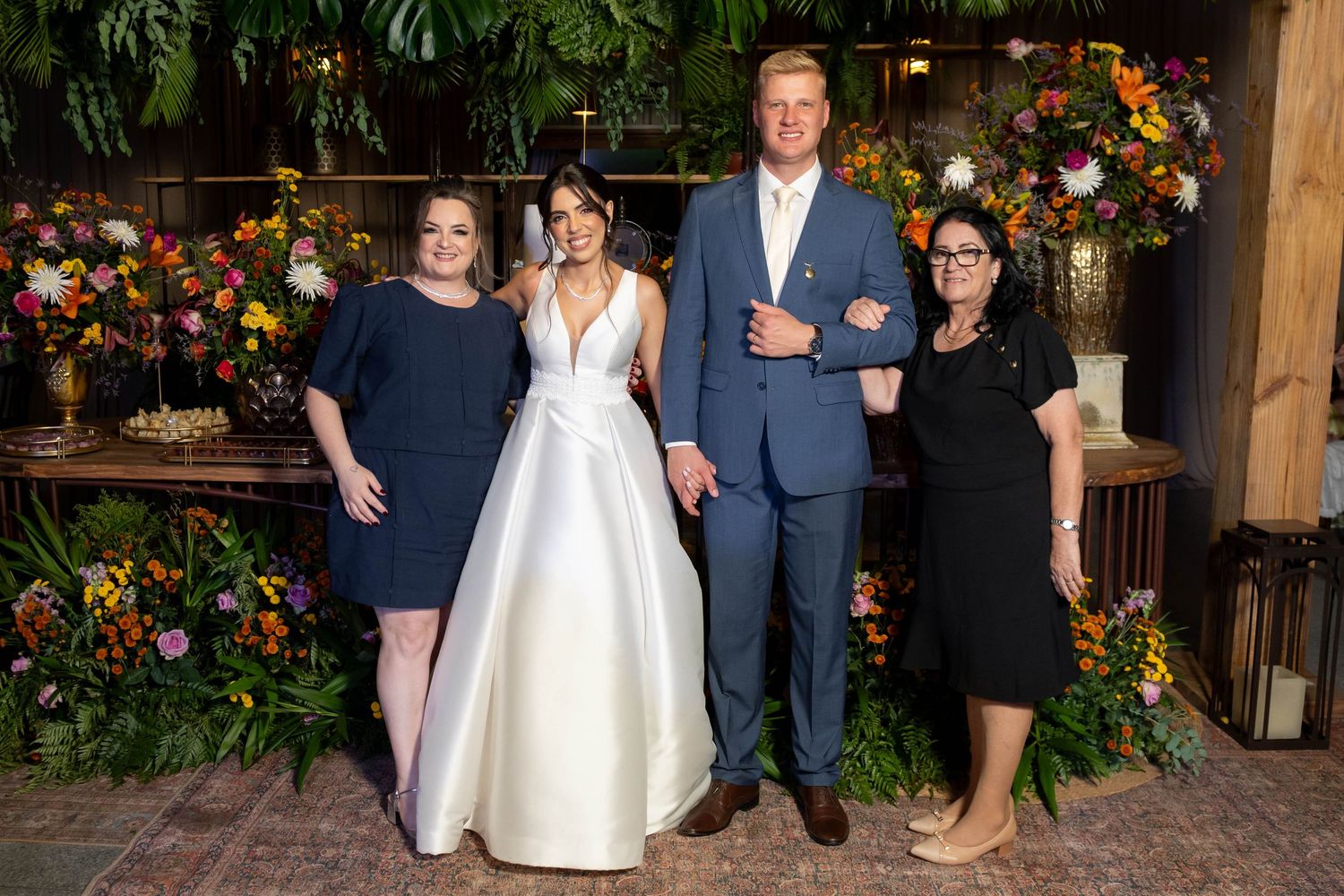 Bride in white gown poses with groom in blue suit and two guests amid colorful floral backdrop.