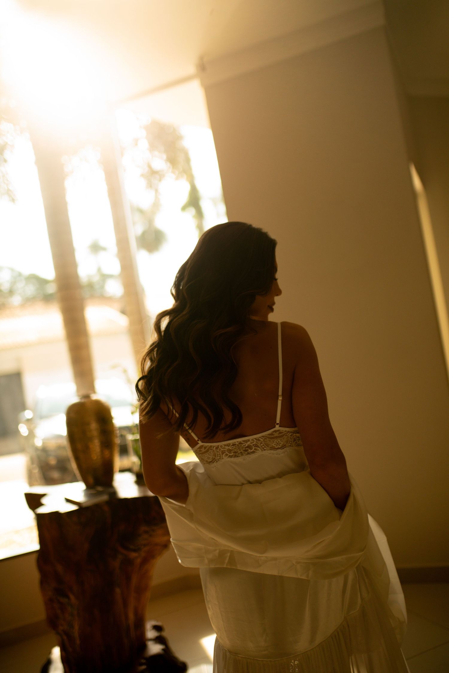 Bride in white wedding dress with lace back stands in golden sunlight near large windows with flowing dark hair.