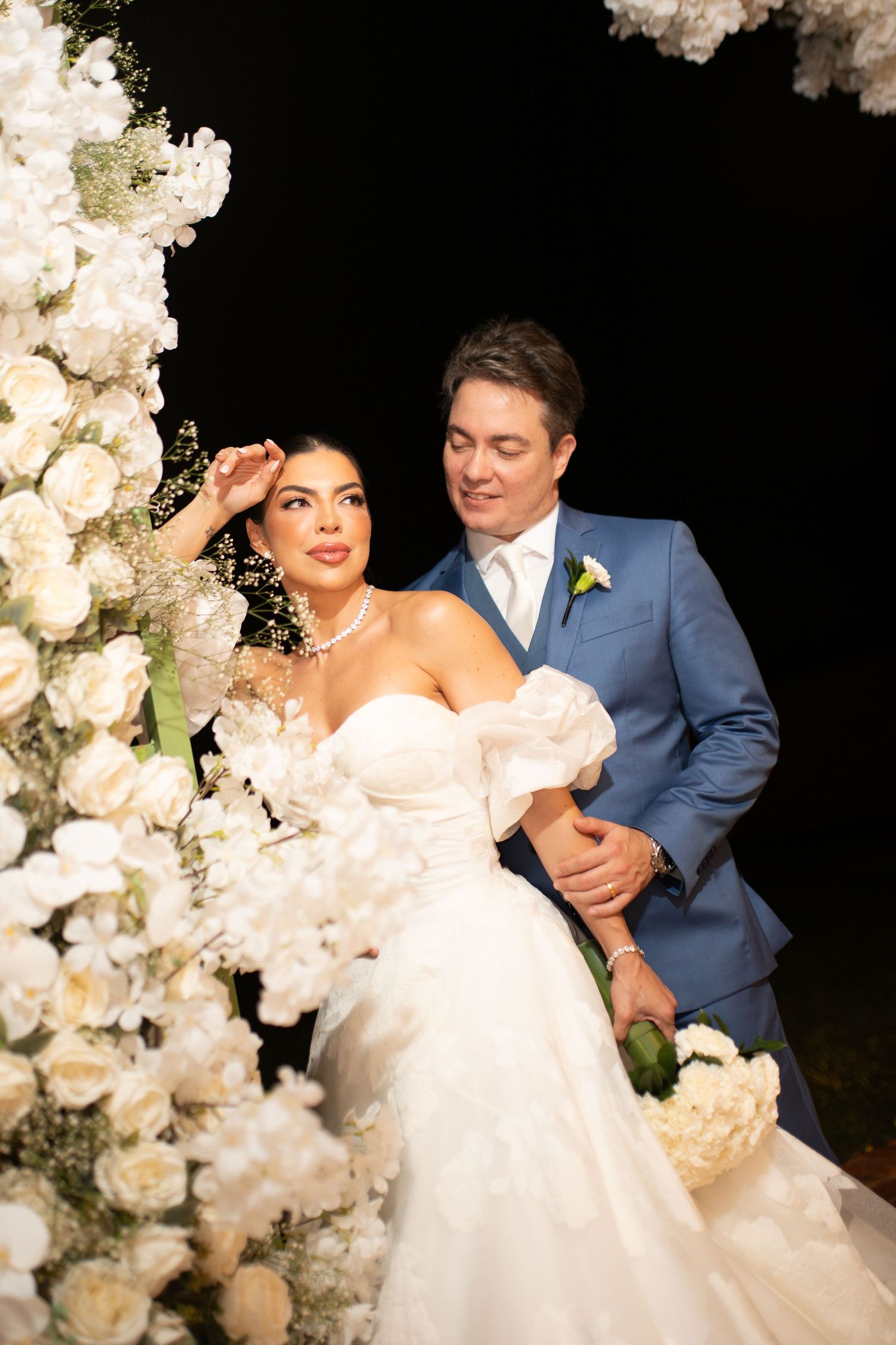 Elegant bride in white off-shoulder gown and groom in blue suit pose together among white floral arrangements at wedding.