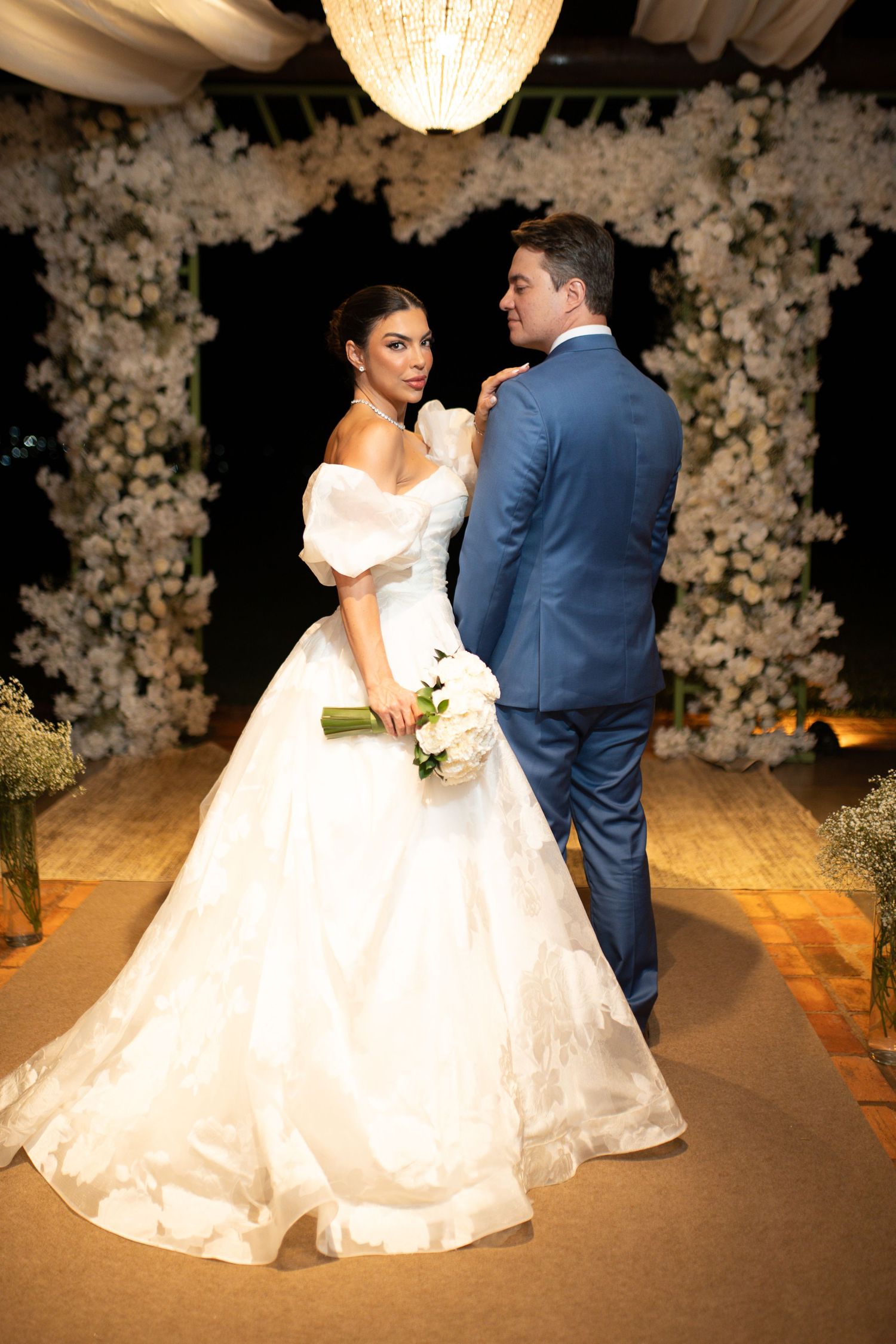 Bride in white ballgown holds bouquet beside groom in blue suit at elegant floral wedding ceremony altar.
