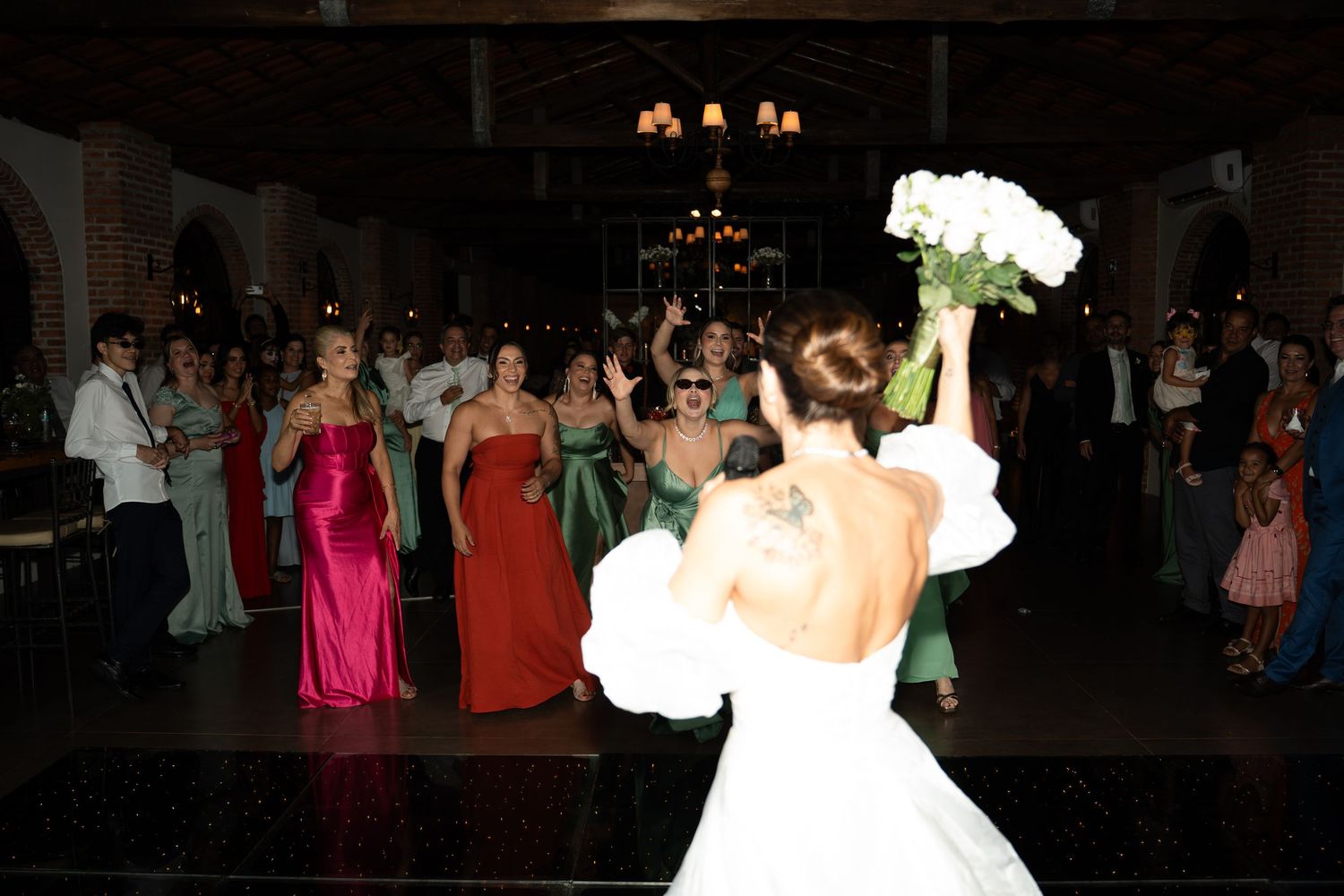 Bride in white gown tossing bouquet of white roses to bridesmaids in colorful gowns at elegant wedding reception.