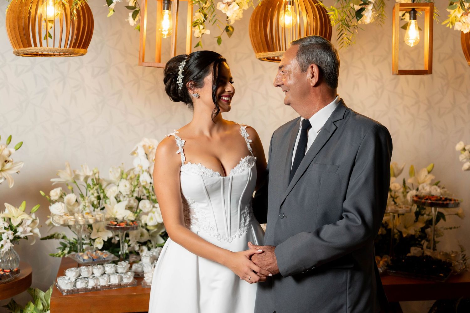 Bride in white wedding dress smiling with older man in gray suit at elegant floral wedding reception.
