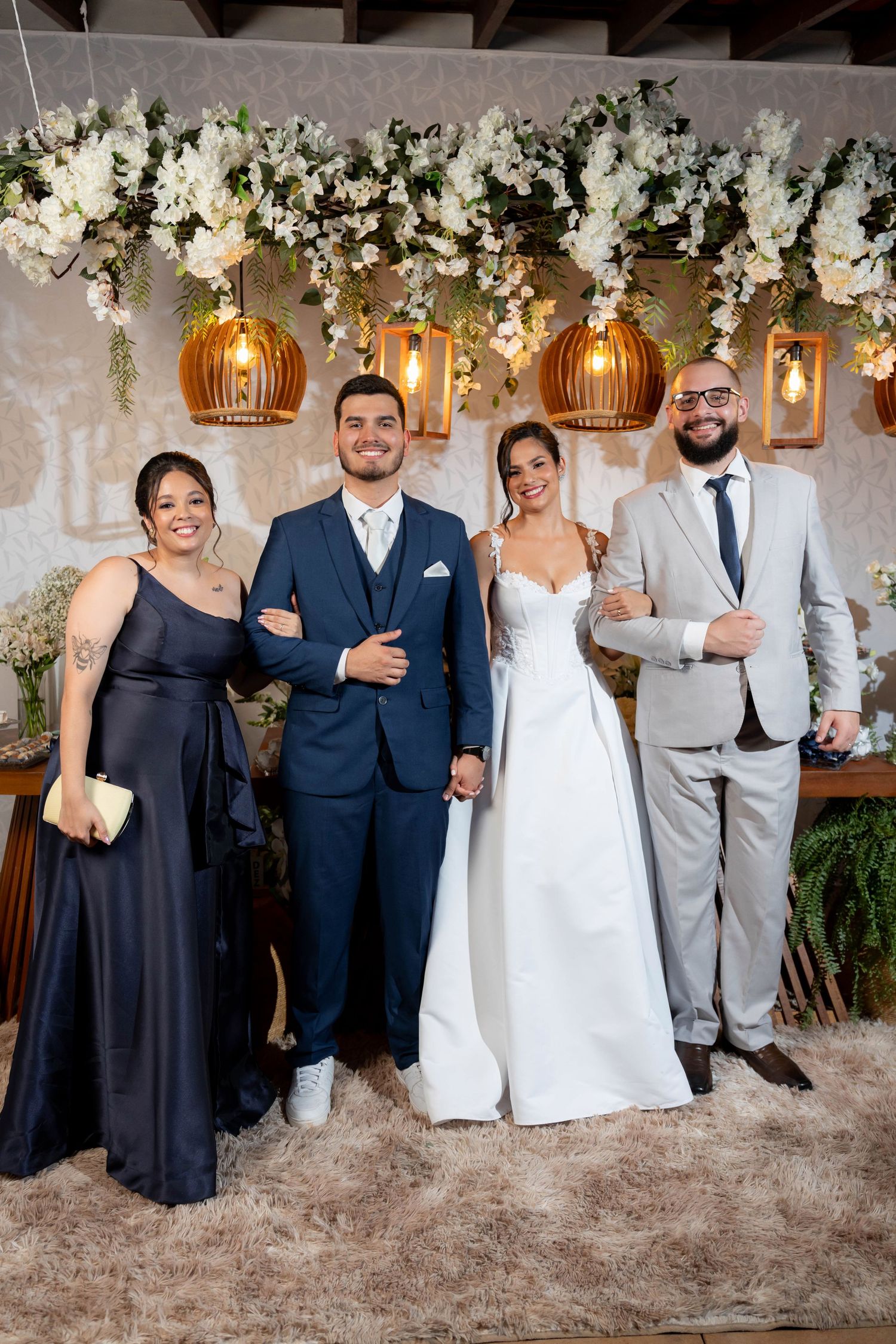 Bride and groom pose with two guests at elegant wedding reception with floral arch and golden pendant lights.