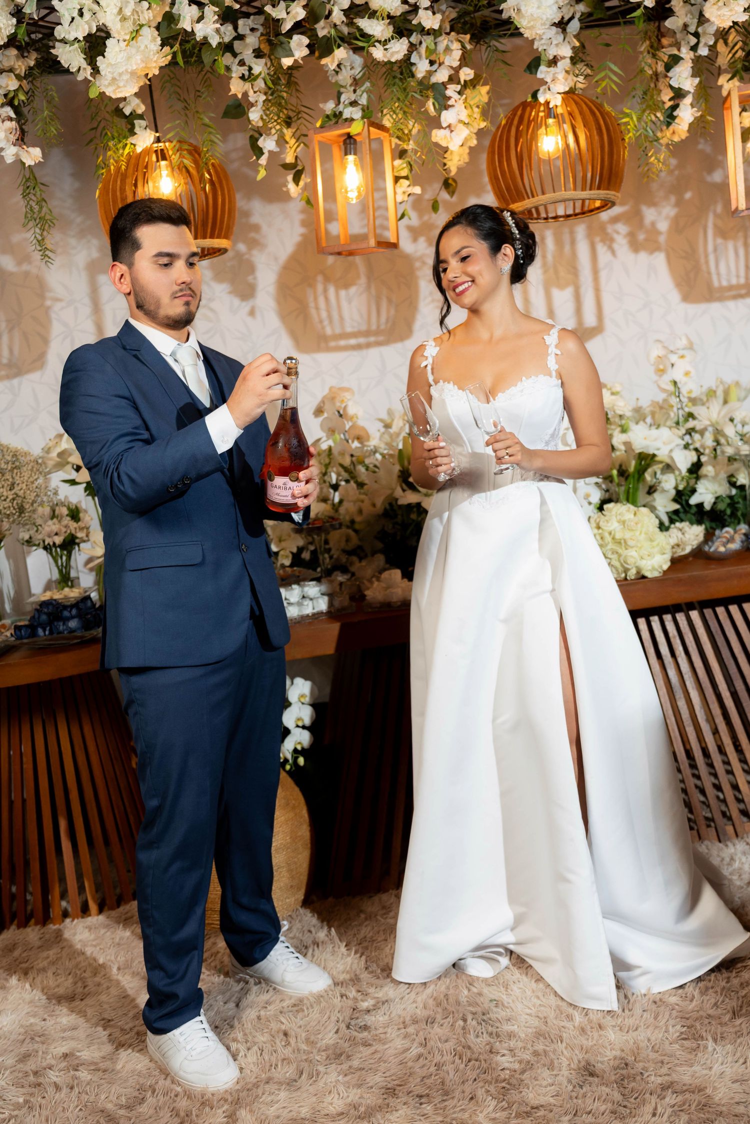 Bride in white gown and groom in navy suit opening champagne bottle at elegant floral wedding reception.