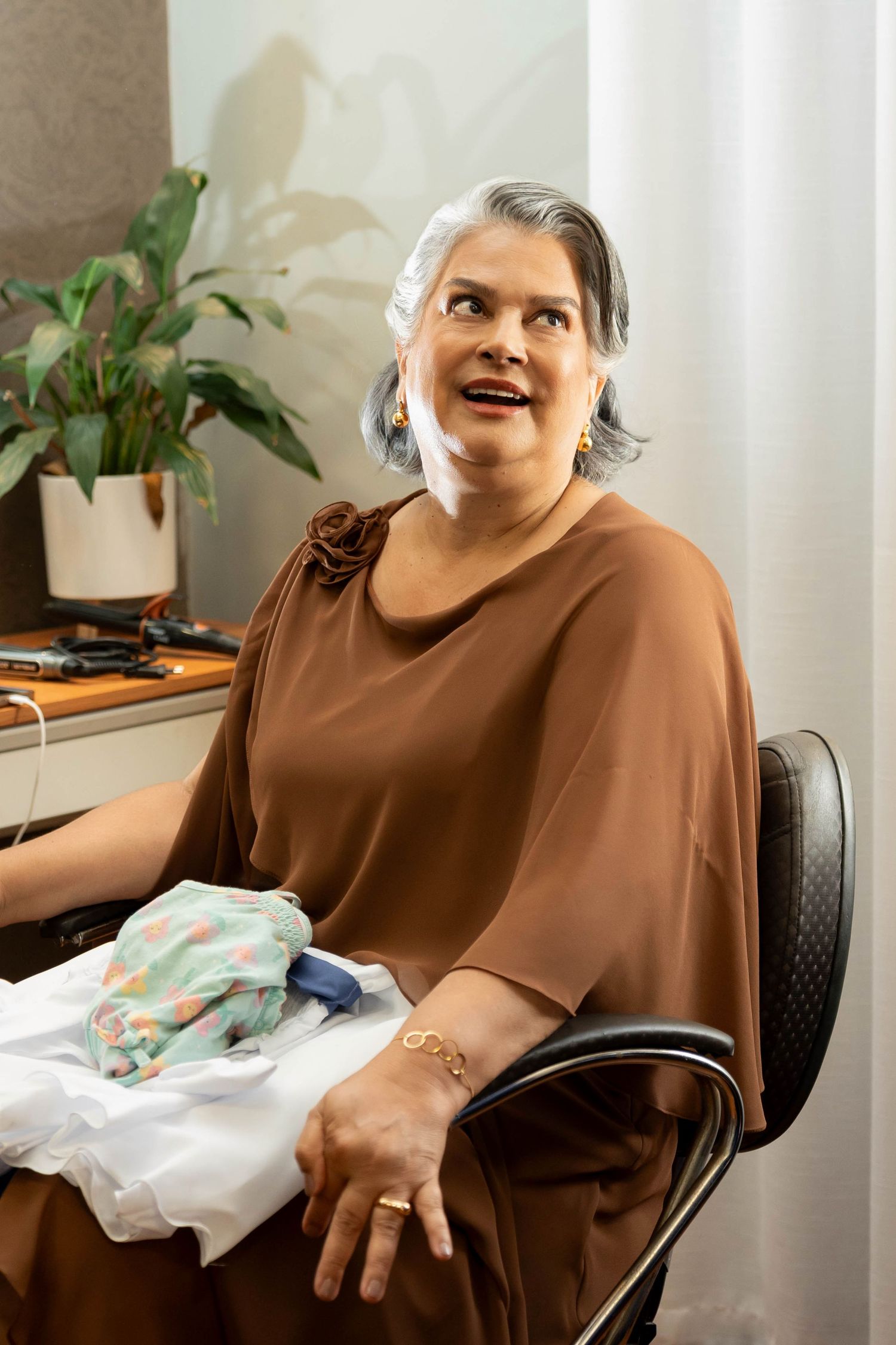 Middle-aged woman with gray hair sits in salon chair wearing brown top, holding fabric samples, smiling upward.