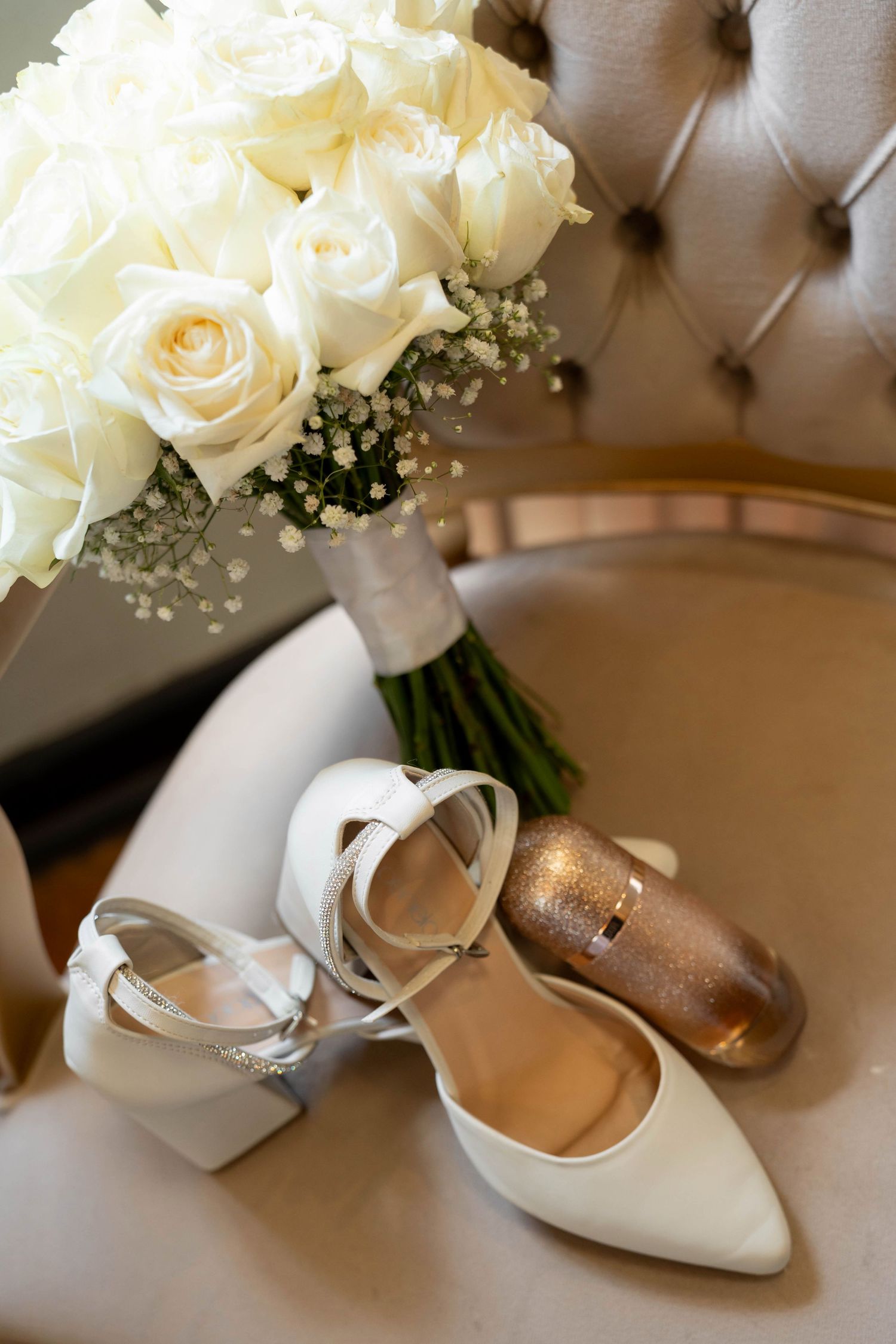 White bridal heels and gold shoes beside a bouquet of white roses on a tufted chair.