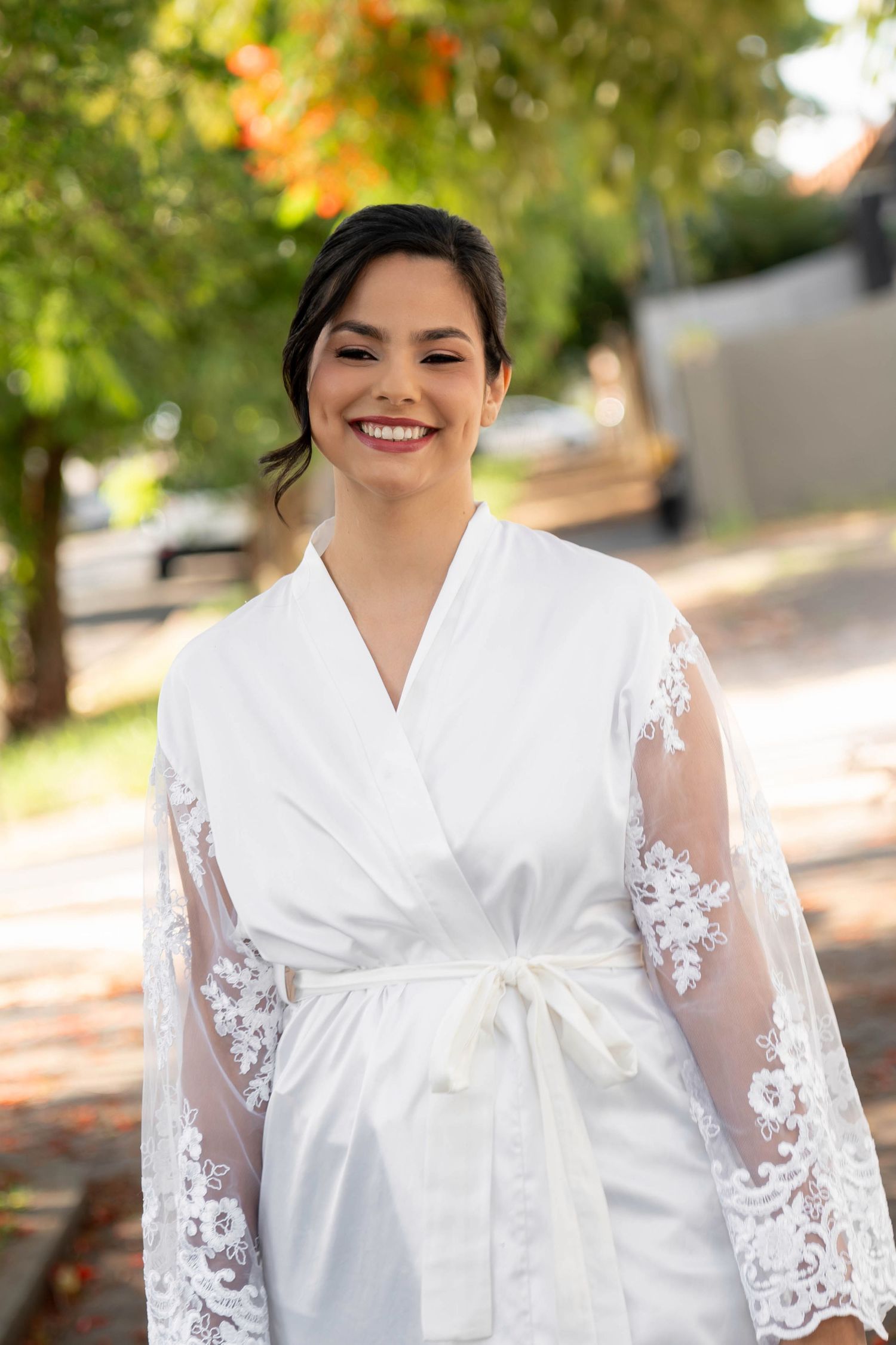 Smiling bride in white lace robe with satin belt posing outdoors among green trees on her wedding day.