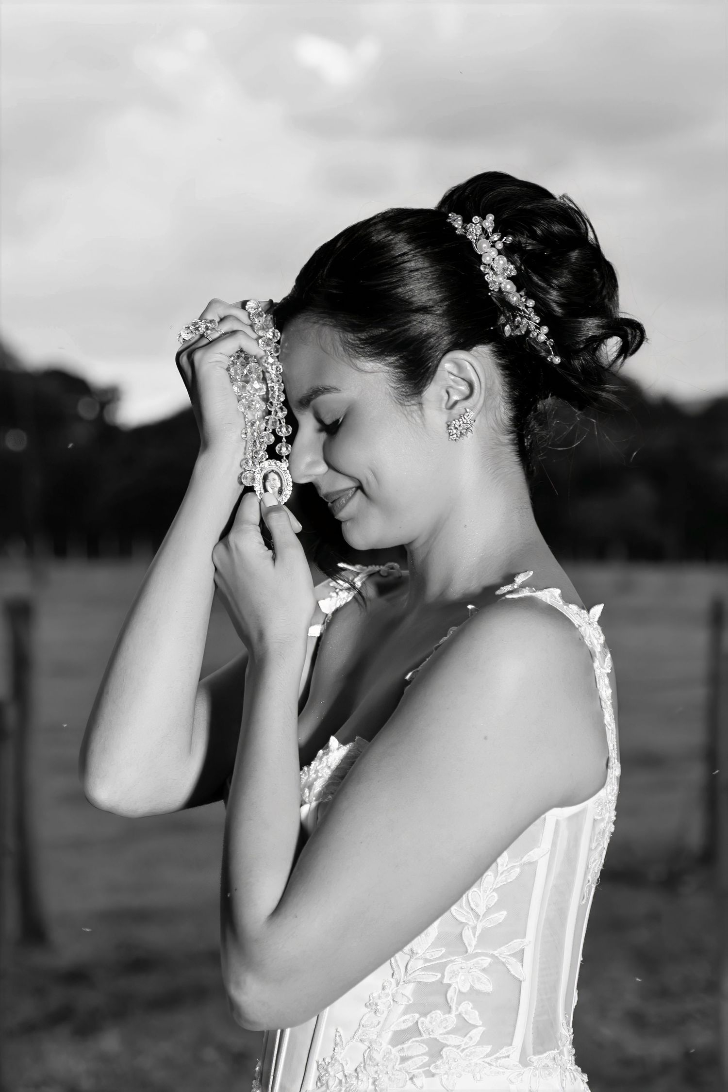 Black and white photo of a smiling bride in lace dress holding jewelry to her forehead outdoors.