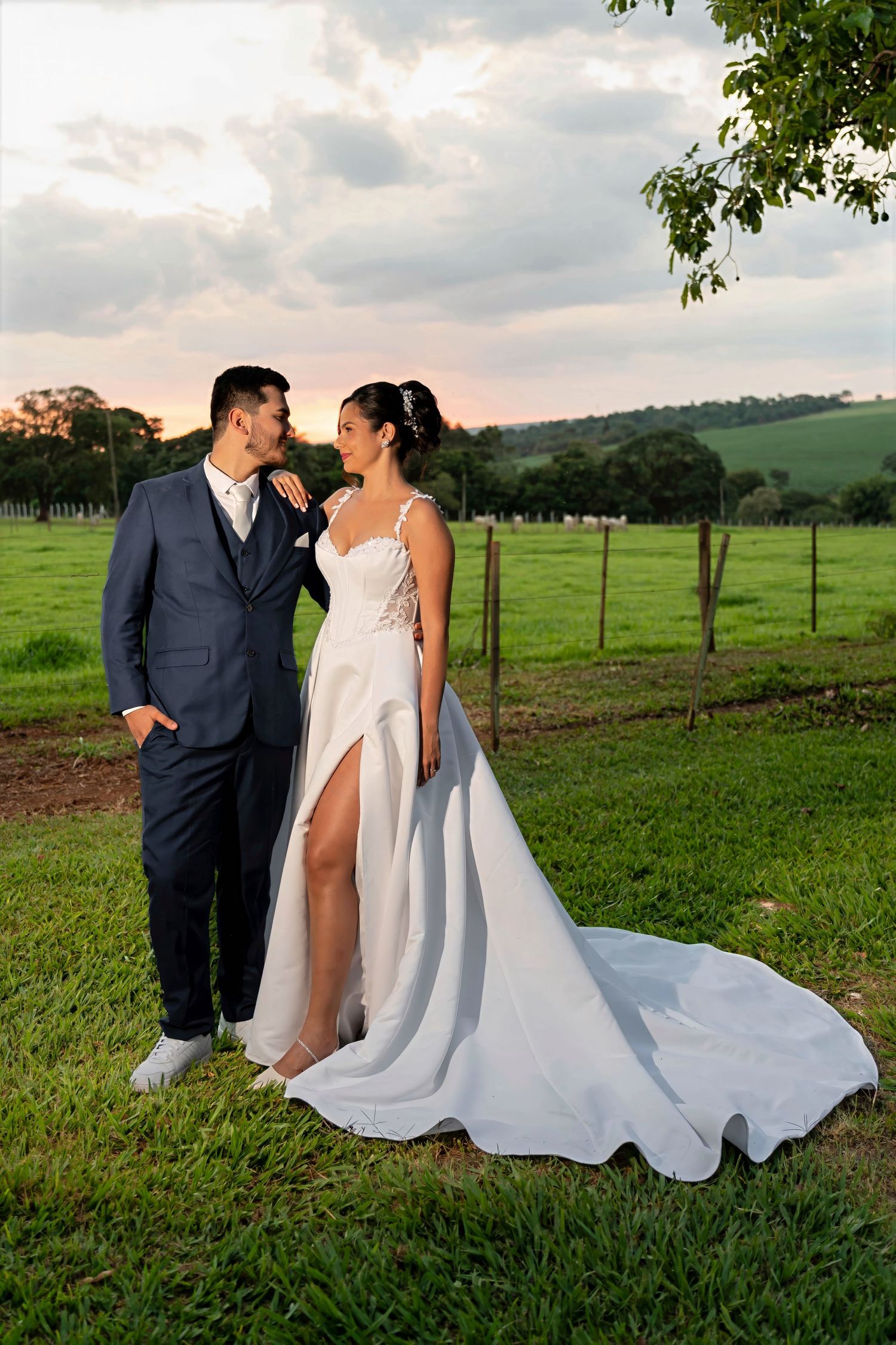 Bride and groom pose romantically at sunset on lush green farm, she in white gown with slit, he in navy suit.