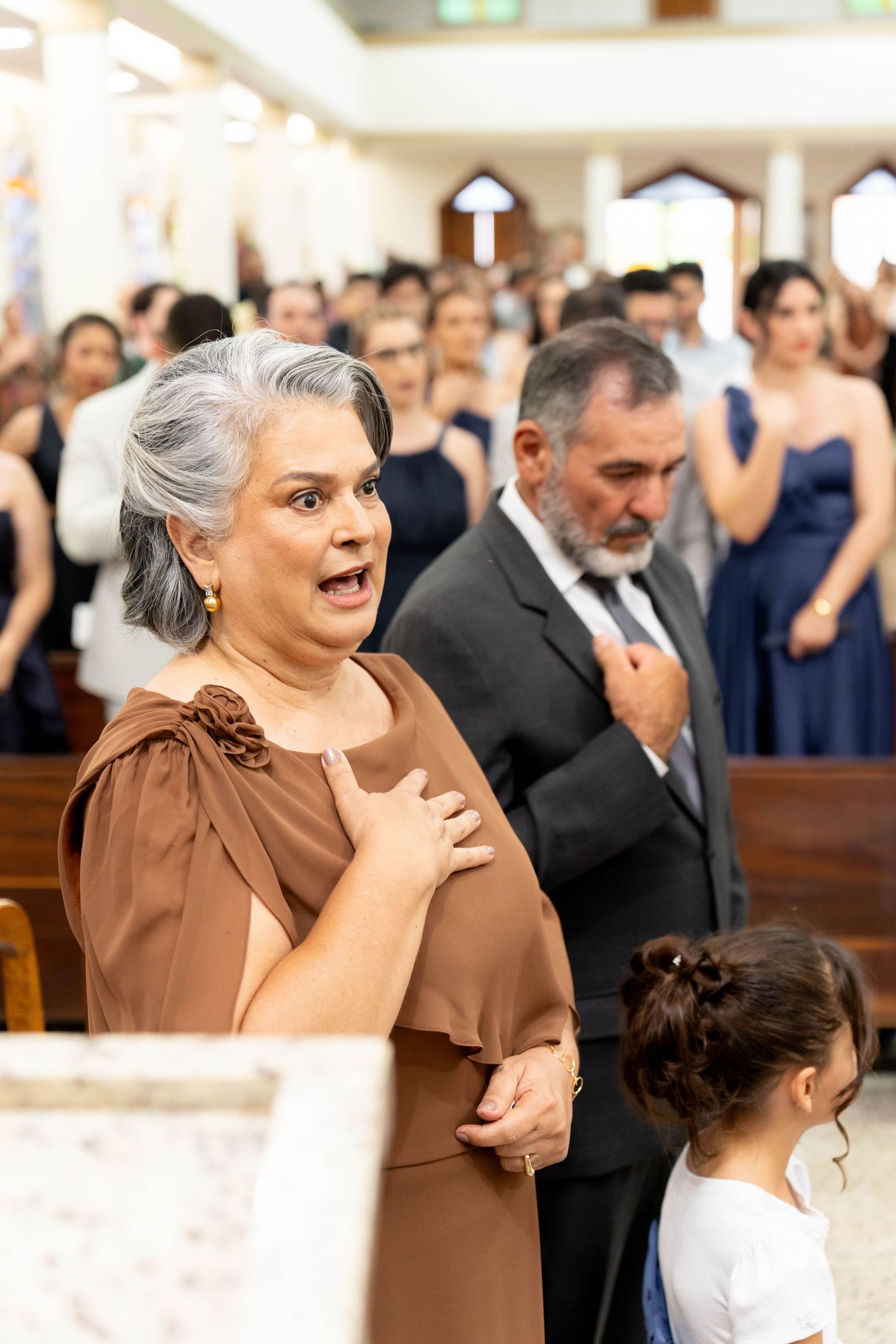 Woman in brown dress at church wedding ceremony, hand on chest, with guests in navy blue attire in background.