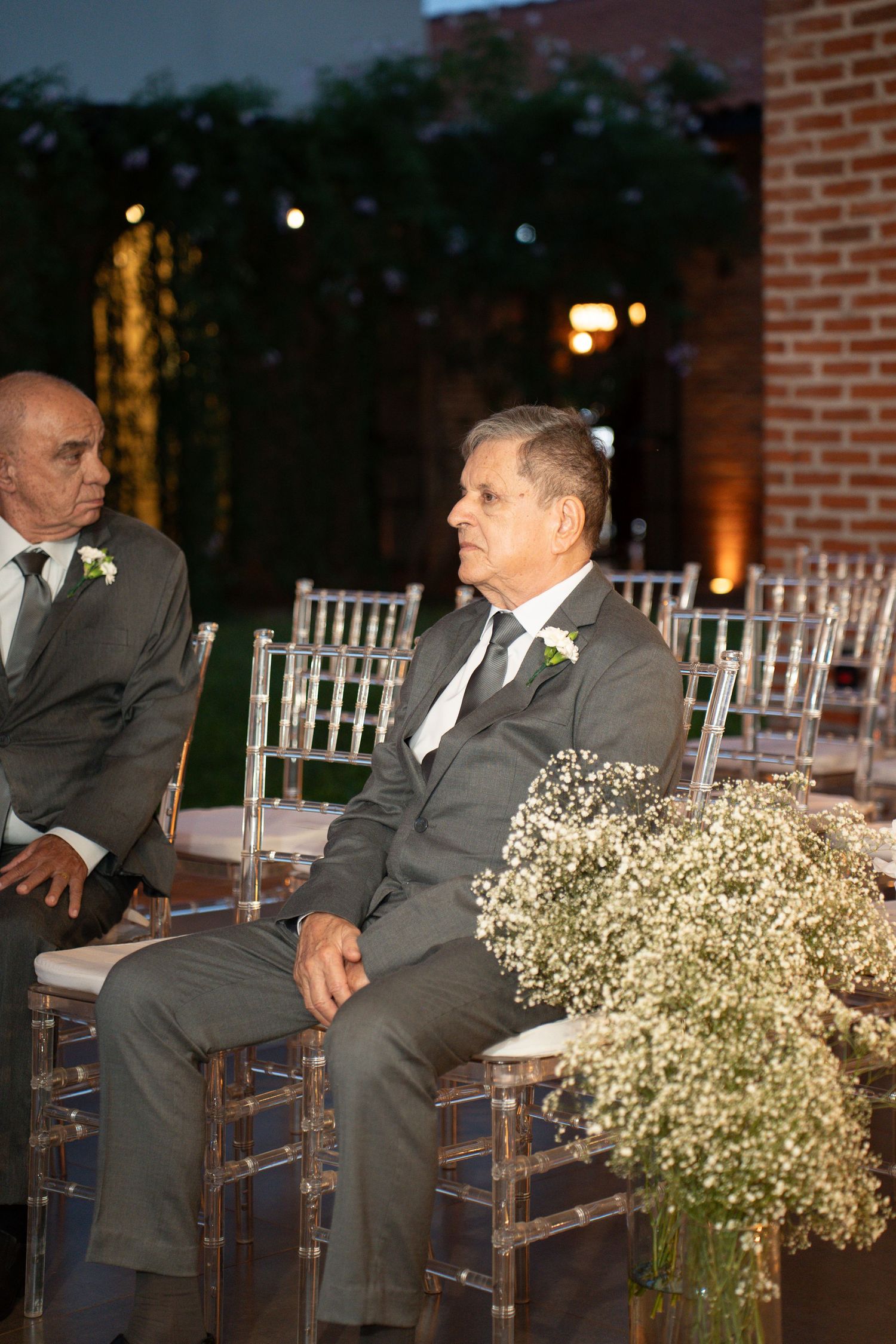 Two elderly men in gray suits with boutonnieres seated at an outdoor evening wedding ceremony.
