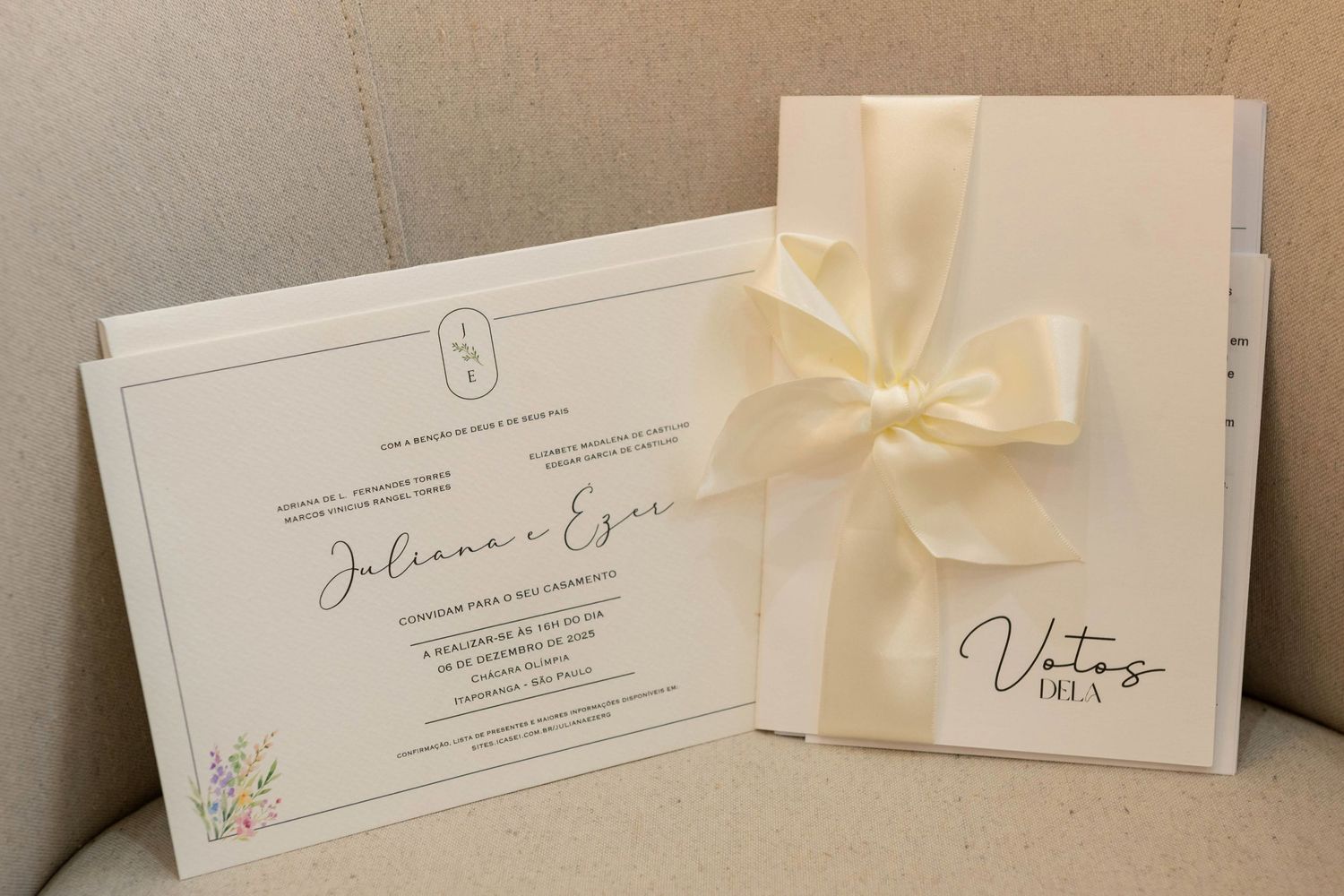 Elegant wedding invitation suite with floral details and a vow booklet tied with a cream satin ribbon on a beige sofa.