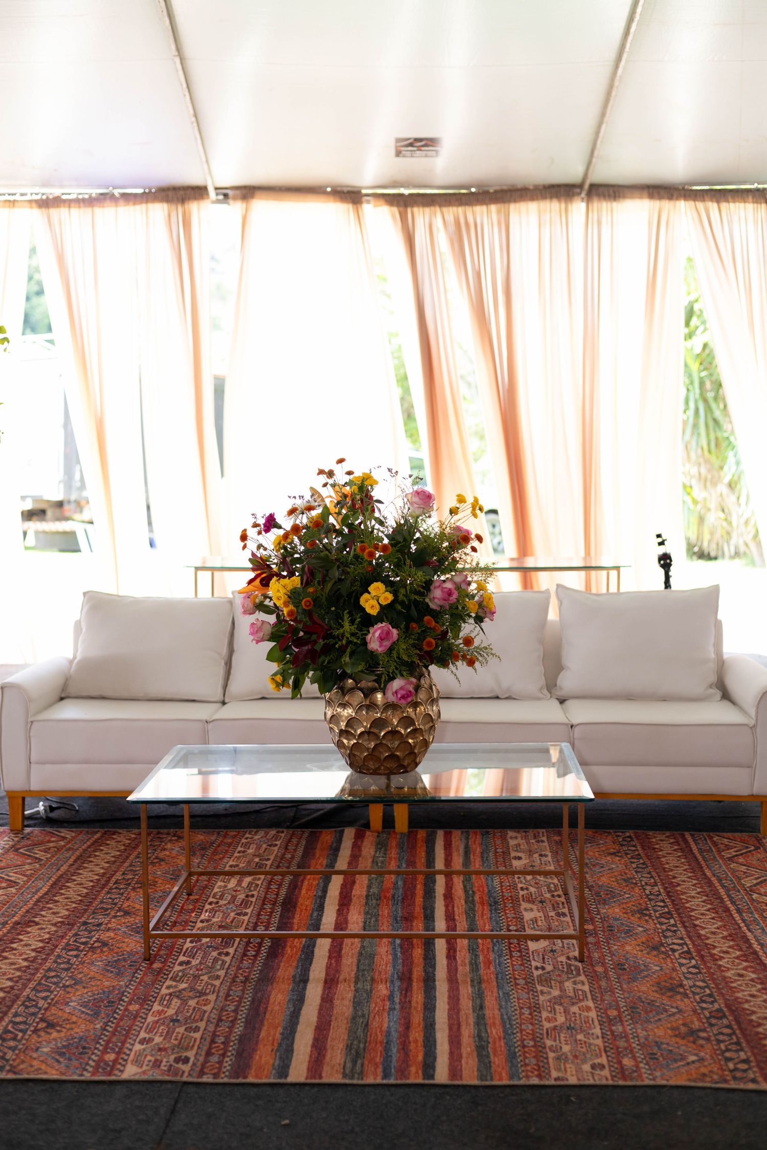 White sofa with colorful floral arrangement on glass coffee table atop striped rug under draped tent.