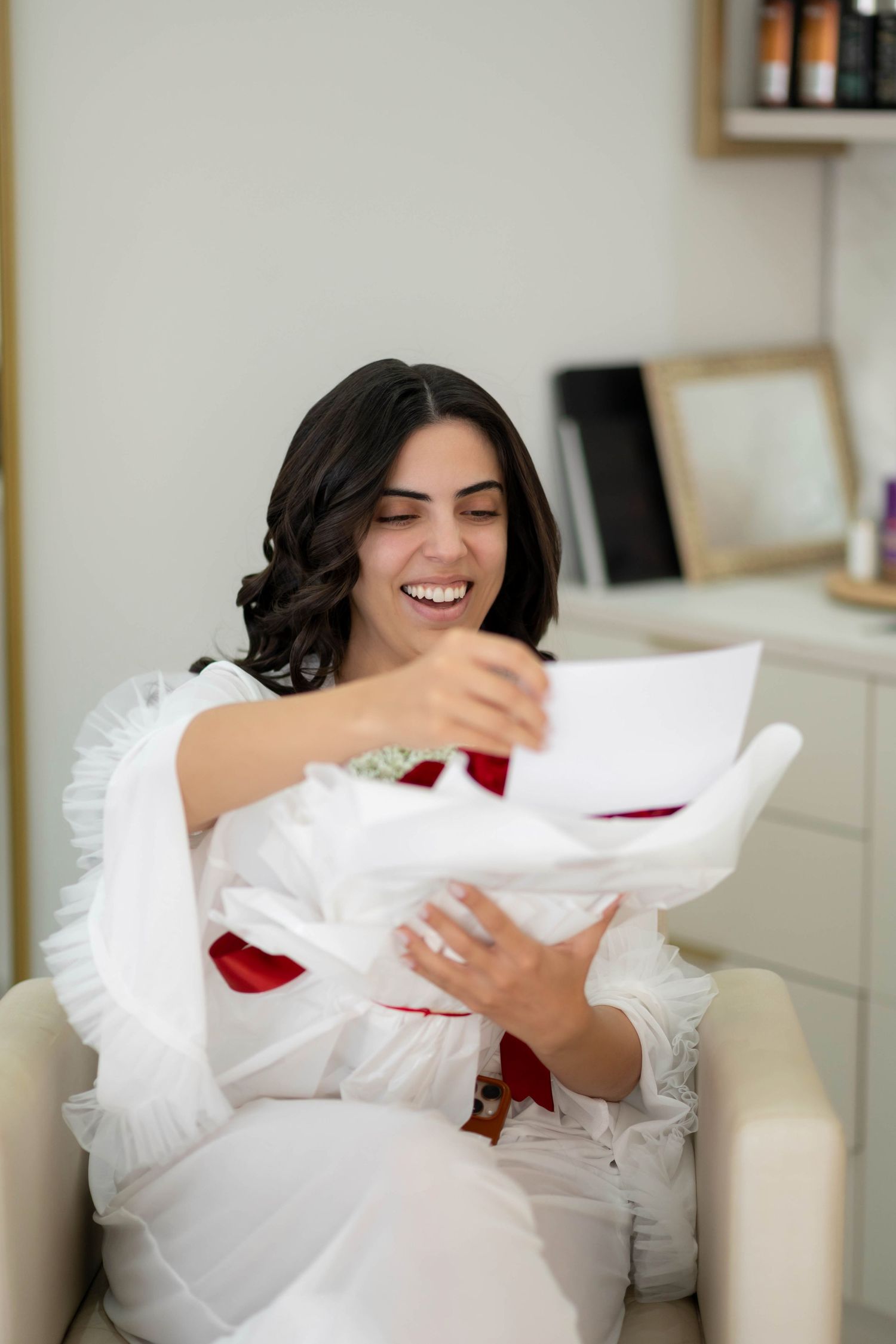 A joyful woman in a white ruffled dress laughs while opening a gift box in a bright, elegant room.