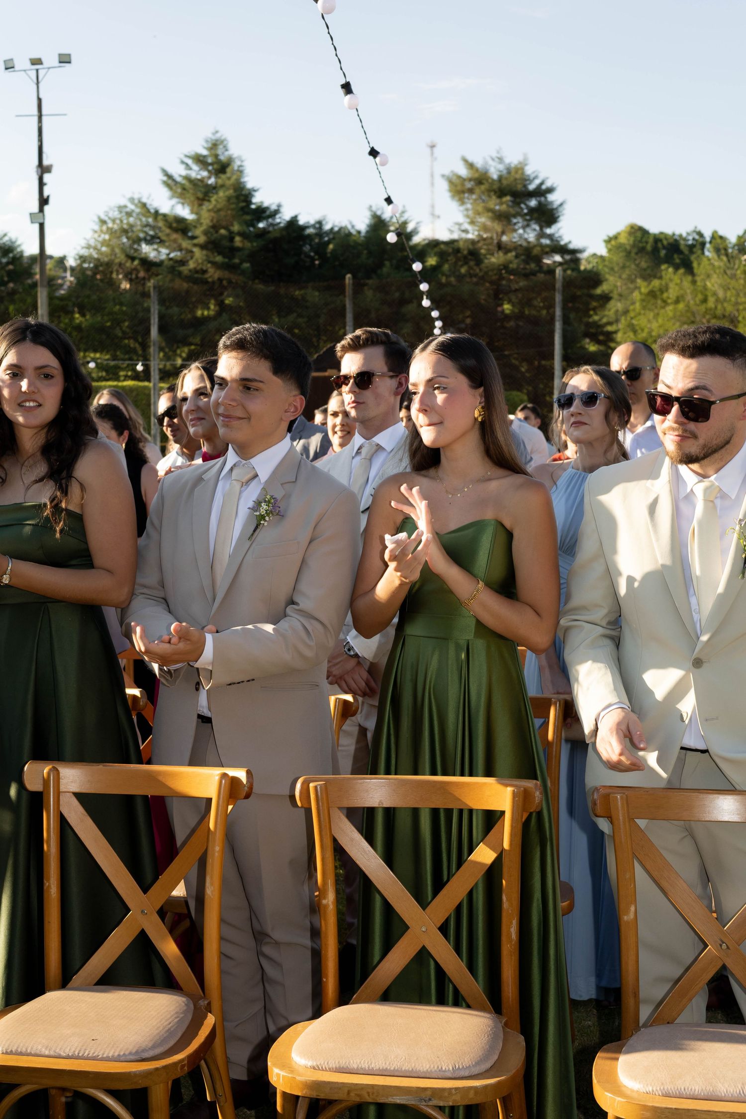 Guests in green dresses and tan suits applaud at an outdoor wedding ceremony with wooden chairs.