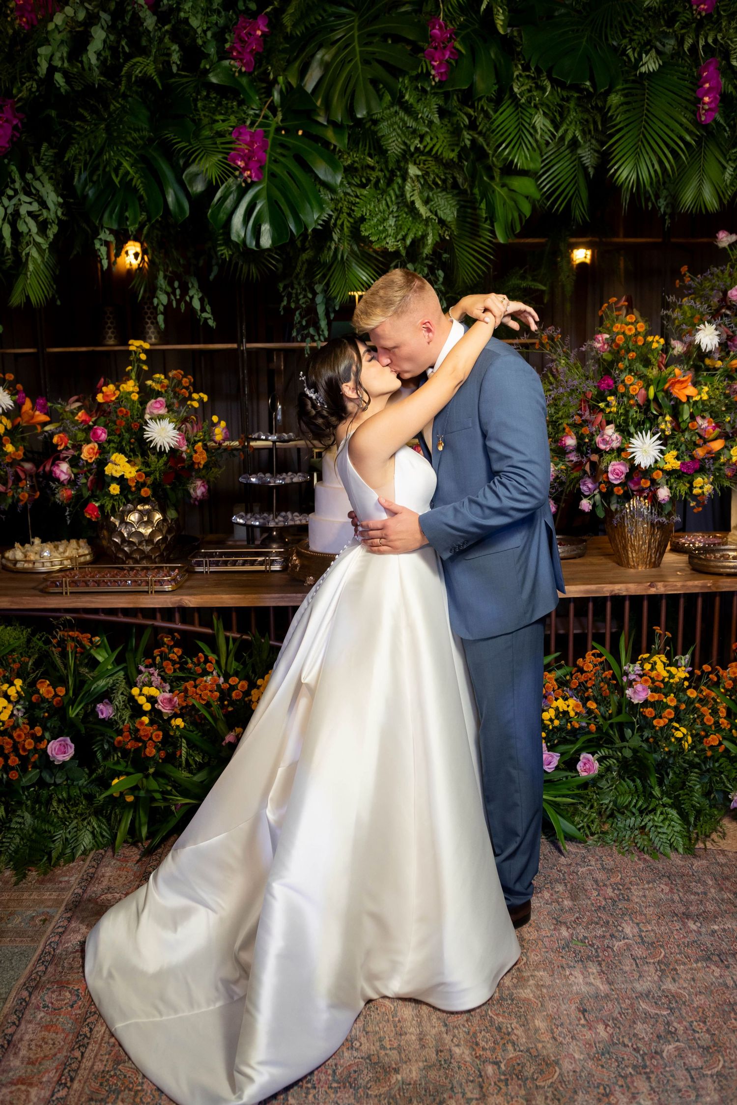 Bride and groom share first dance embrace surrounded by lush tropical greenery and colorful floral arrangements at wedding reception.