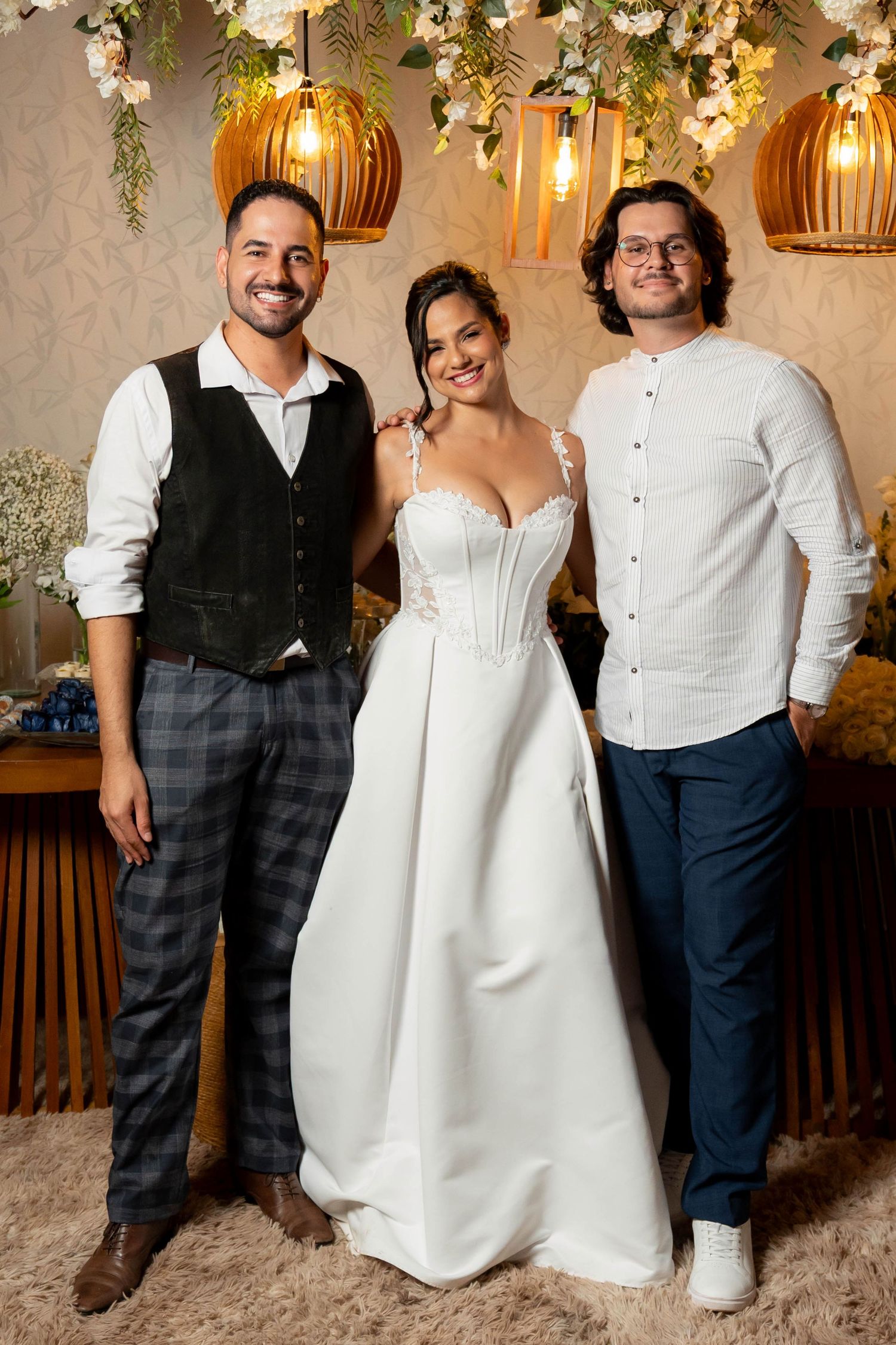 Bride in white gown poses with two men in elegant wedding venue decorated with floral arrangements and warm pendant lights.