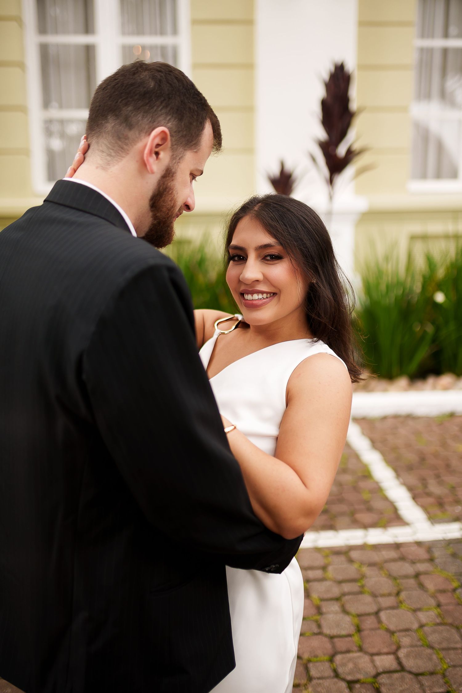 Smiling bride in white dress dancing with groom in black suit outdoors near yellow building.